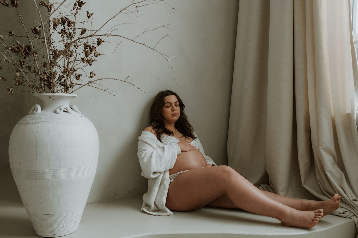 Pregnant woman in white robe sitting on floor beside tall white ceramic vase with dried branches.