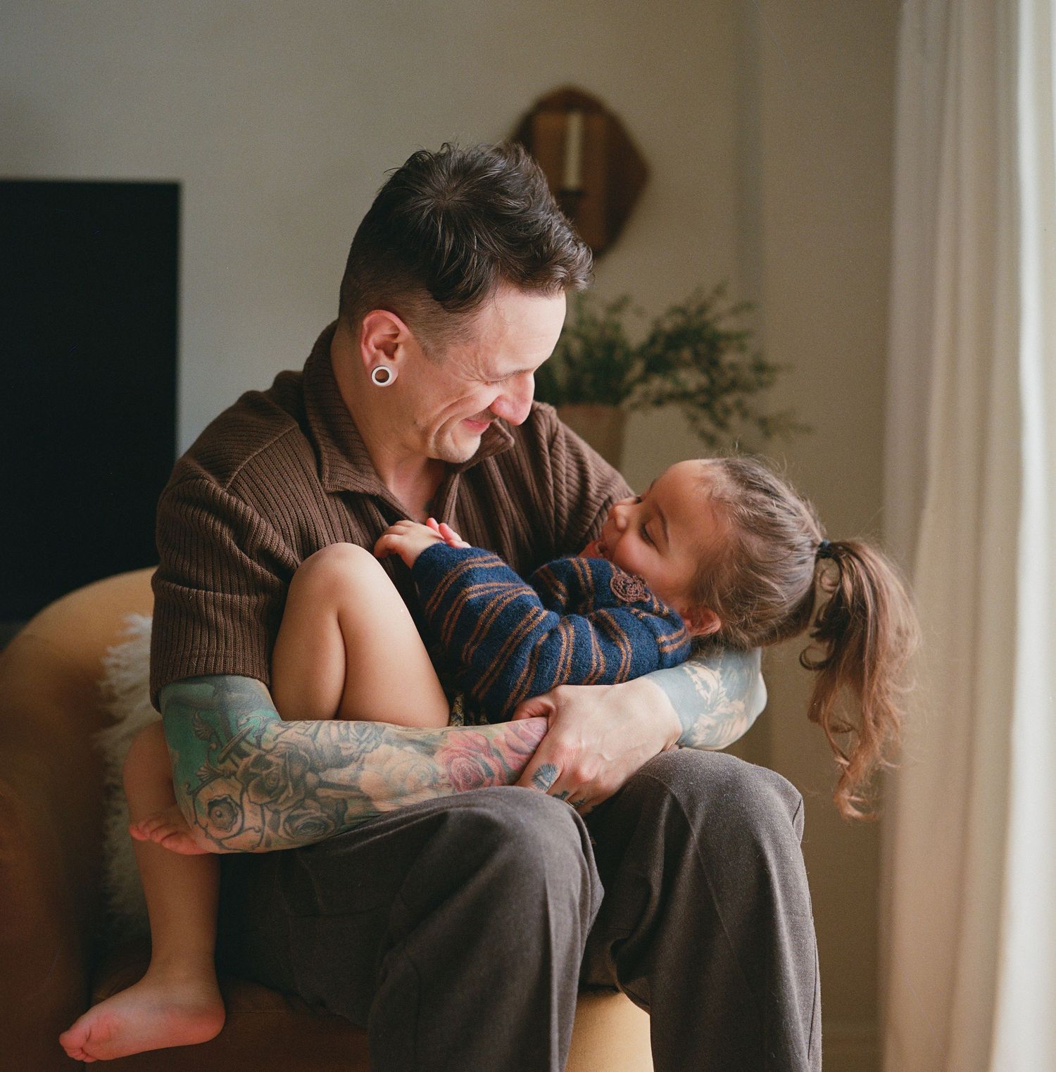 A tattooed man sits in a chair by a window, smiling warmly while holding a laughing young child.