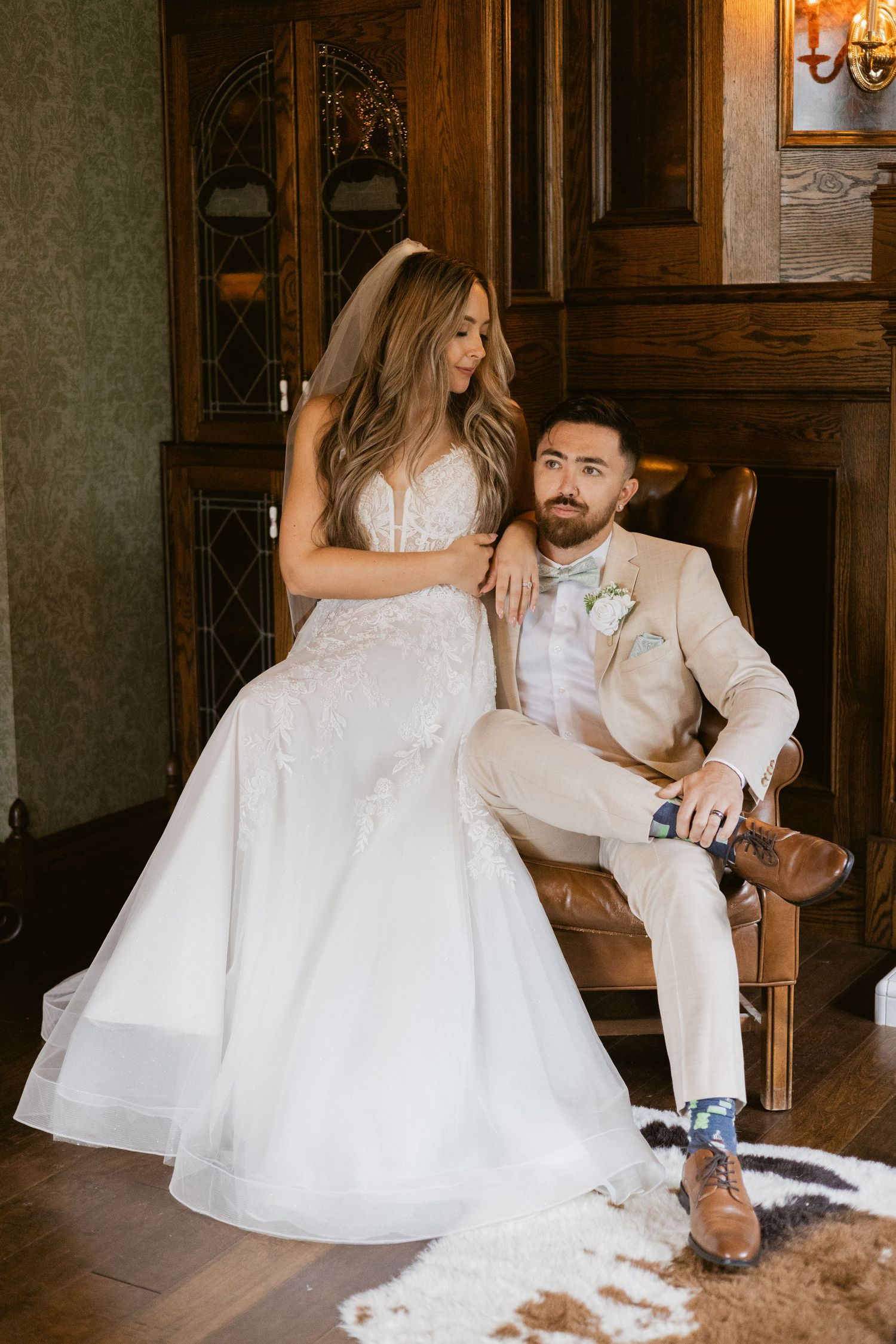 Bride in fitted lace gown stands beside seated groom in beige suit in an elegant wood-paneled room.