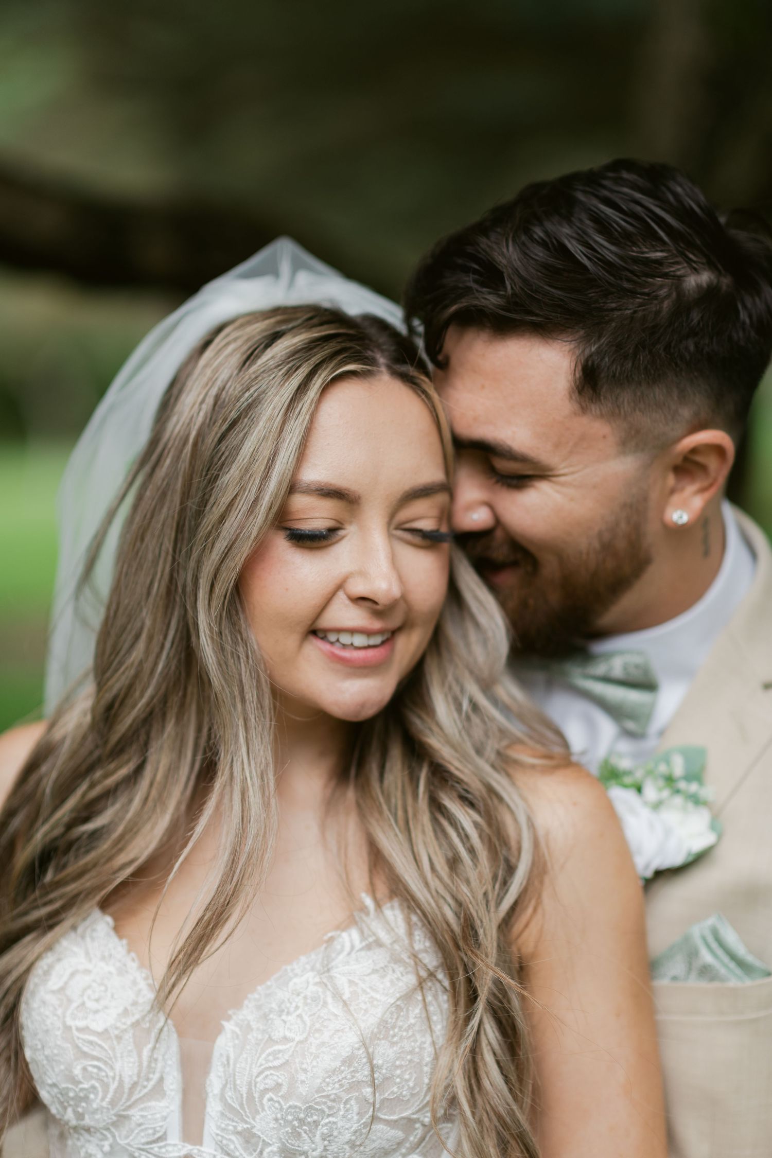 Close-up of smiling bride in lace gown with veil as groom nuzzles her cheek tenderly.