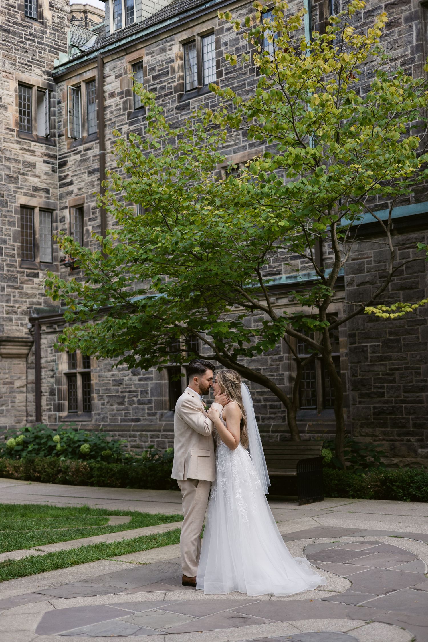Bride and groom share an intimate moment under a green tree beside a historic stone building on their wedding day.