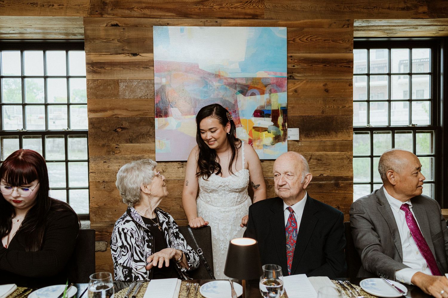 Bride in white dress standing beside elderly guests at a wedding reception with colorful artwork behind.