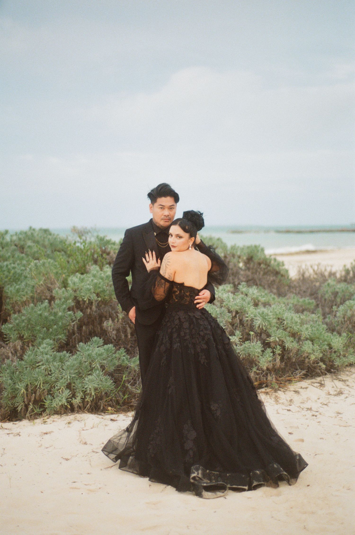 Bride in black floral ballgown leaning against groom, ocean and coastal shrubs backdrop, elegant beach wedding.