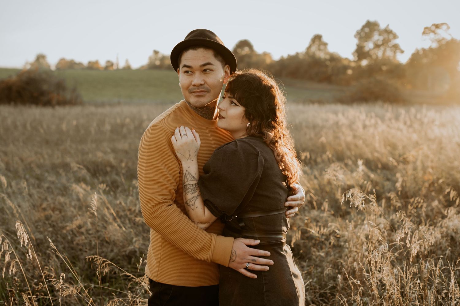 Couple posing at golden hour in autumn field, woman hugging tattooed man in mustard sweater wearing black hat.