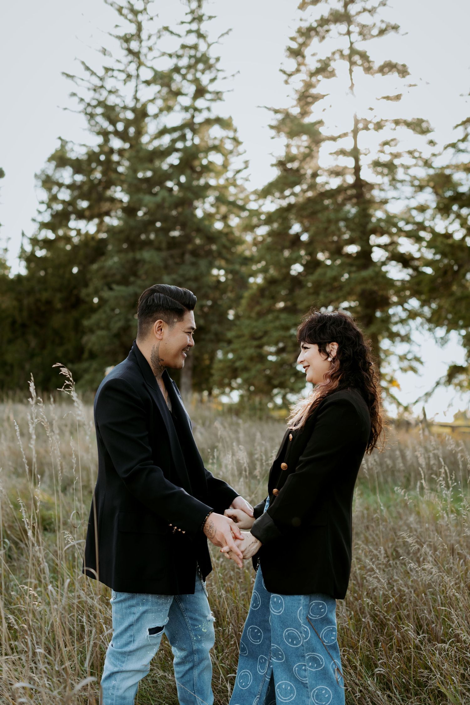 A smiling couple holding hands facing each other in a field with tall evergreen trees behind.