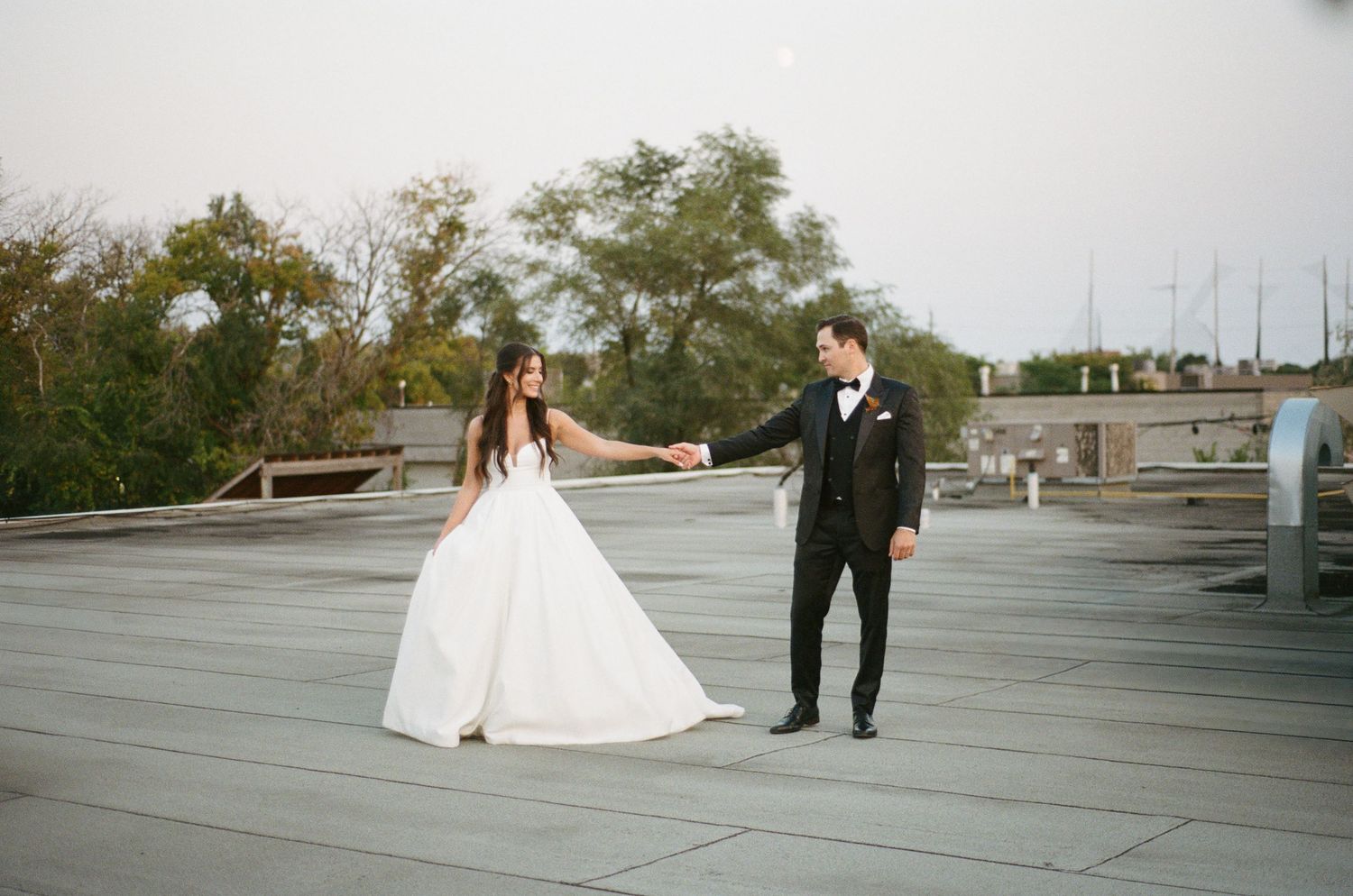 Bride and groom hold hands on rooftop with city trees and overcast sky in background.