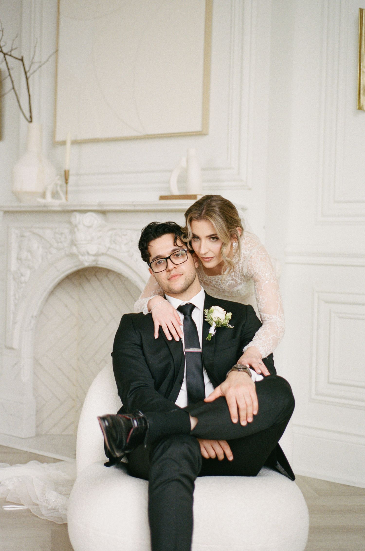Elegant couple posing in white room, groom in black suit seated in chair with bride in lace wedding dress embracing him.