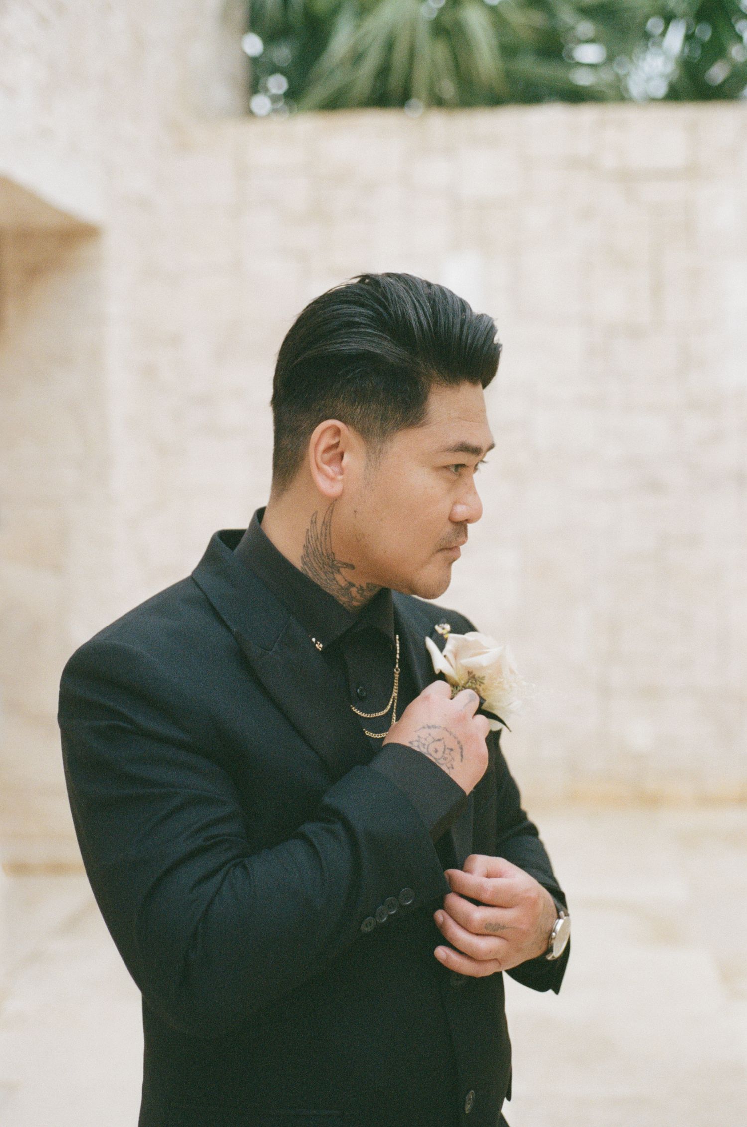 Groom with neck tattoos in black suit holding white flower boutonniere against stone wall.