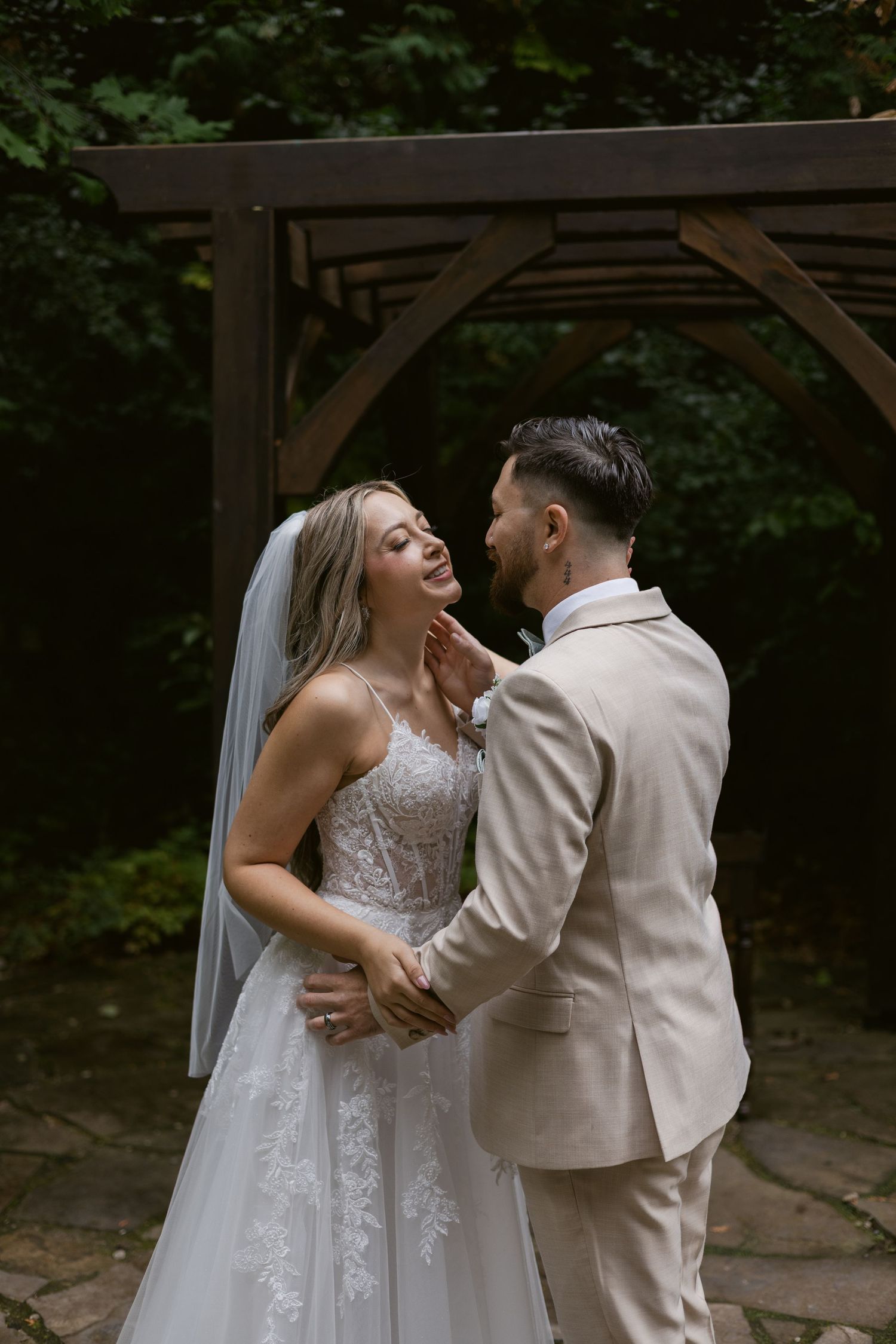 Groom tenderly touching brides face as they share romantic moment under outdoor wooden arch.
