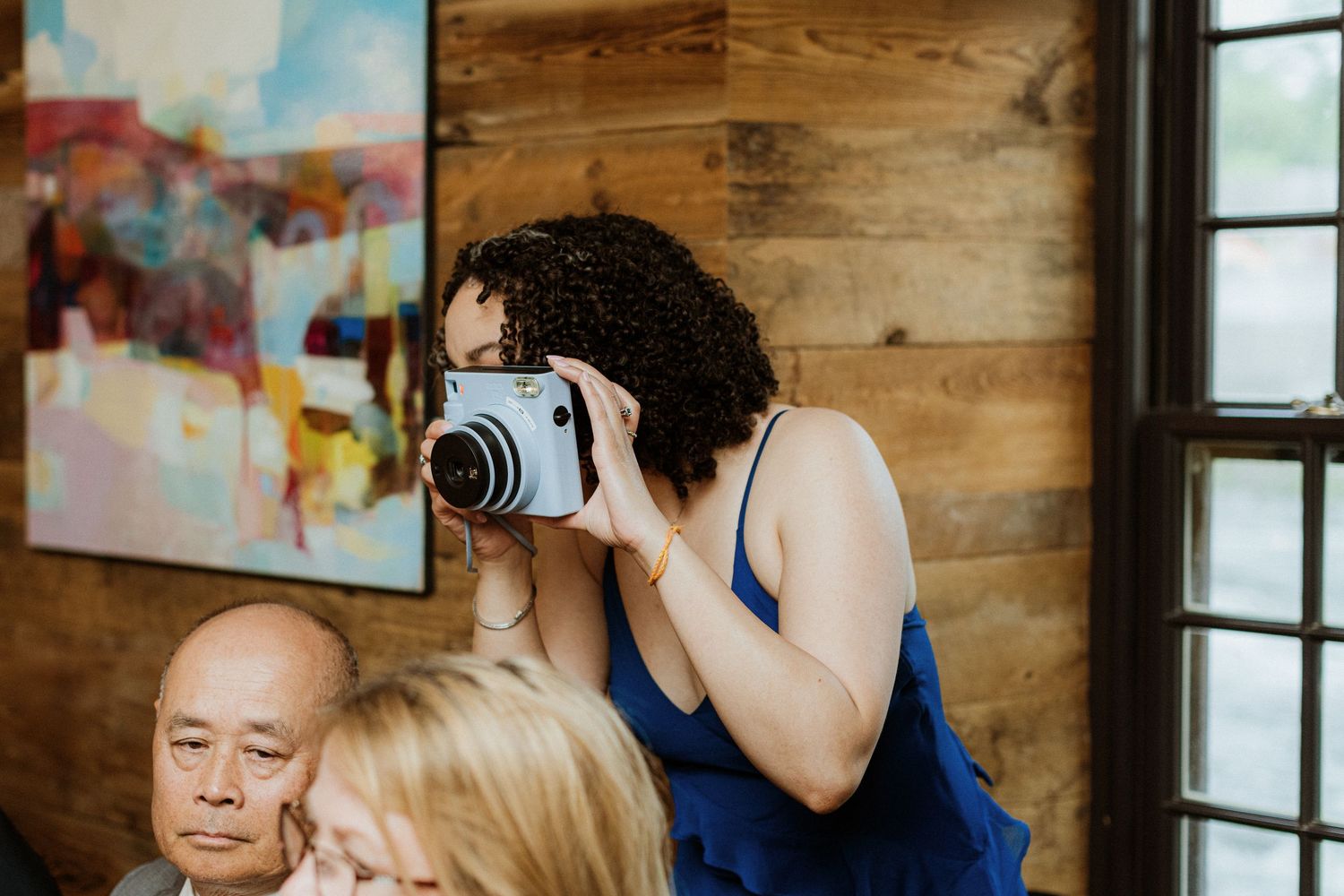 Guest in blue dress photographs wedding reception with instant camera near wooden wall.