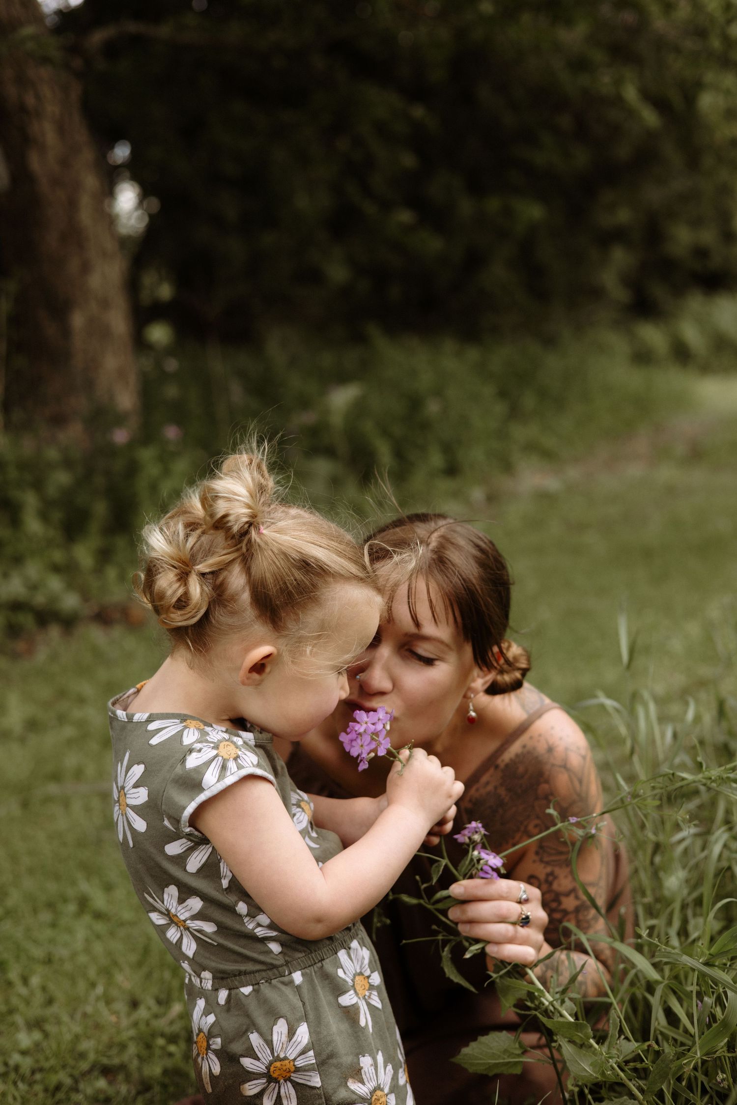 A tattooed mother and young daughter share a tender moment smelling purple wildflowers together in a garden.