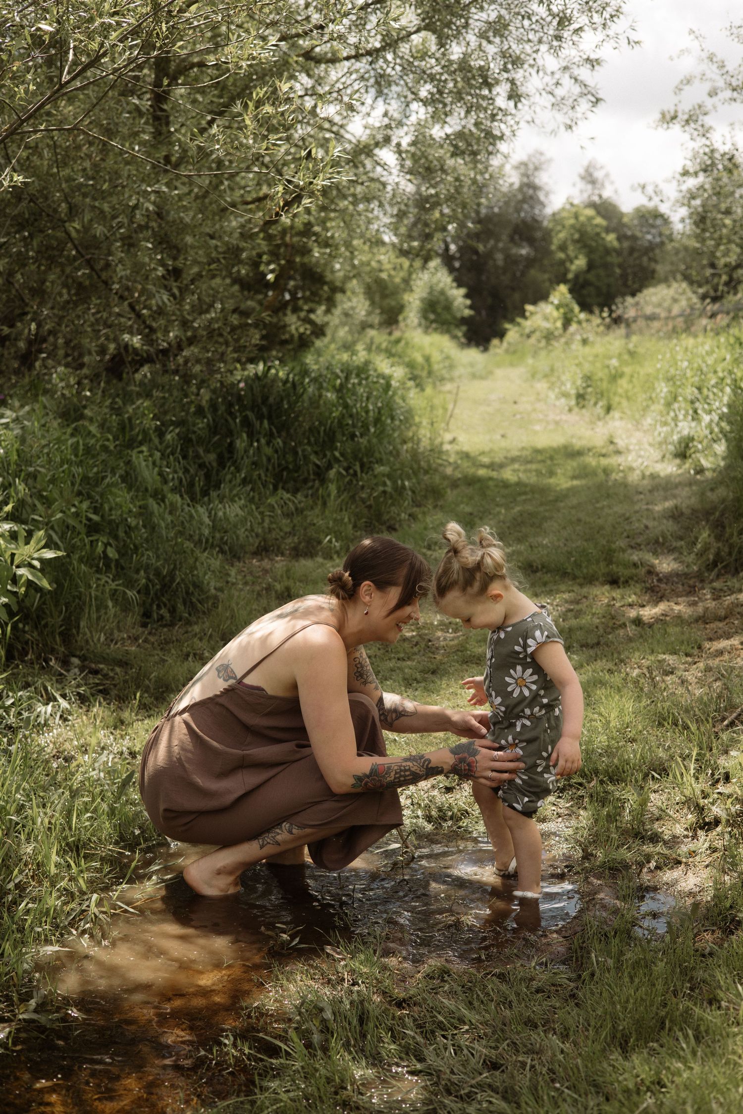 Tattooed mother kneeling in stream helping steady toddler daughter in daisy outfit in a lush green outdoor setting.