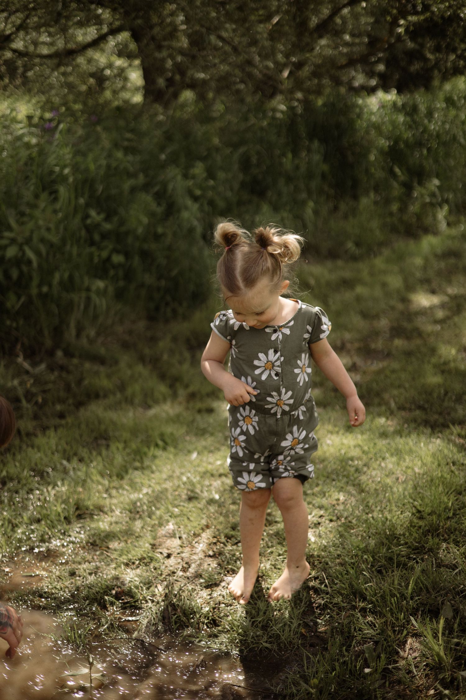 Barefoot toddler girl in a daisy print romper walking on a grassy path in a sunlit green woodland area.