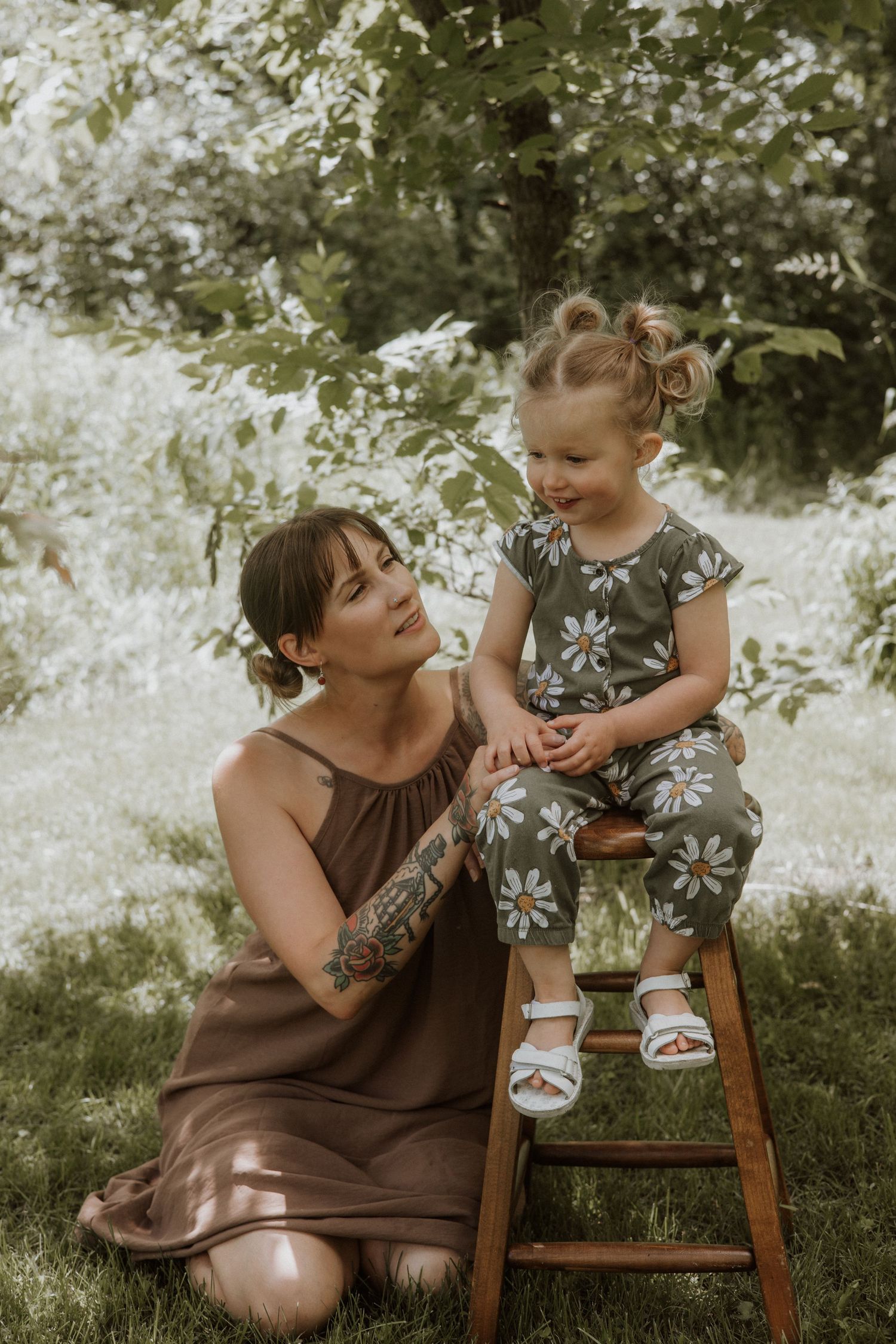 Tattooed mother in brown dress kneeling beside toddler in daisy outfit sitting on wooden stool.