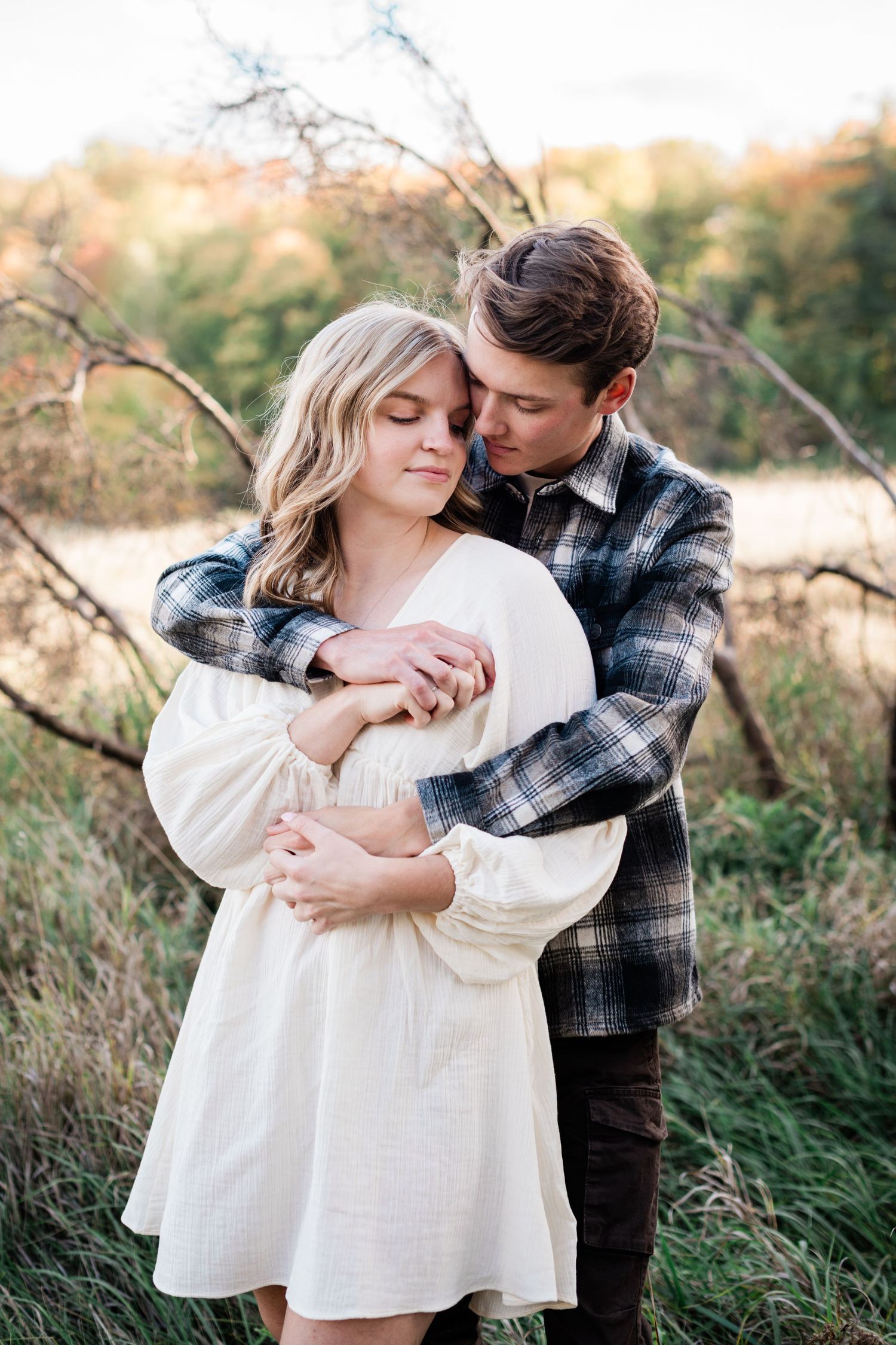 Tender couple embracing in autumn field, man in plaid shirt hugging woman in white dress.