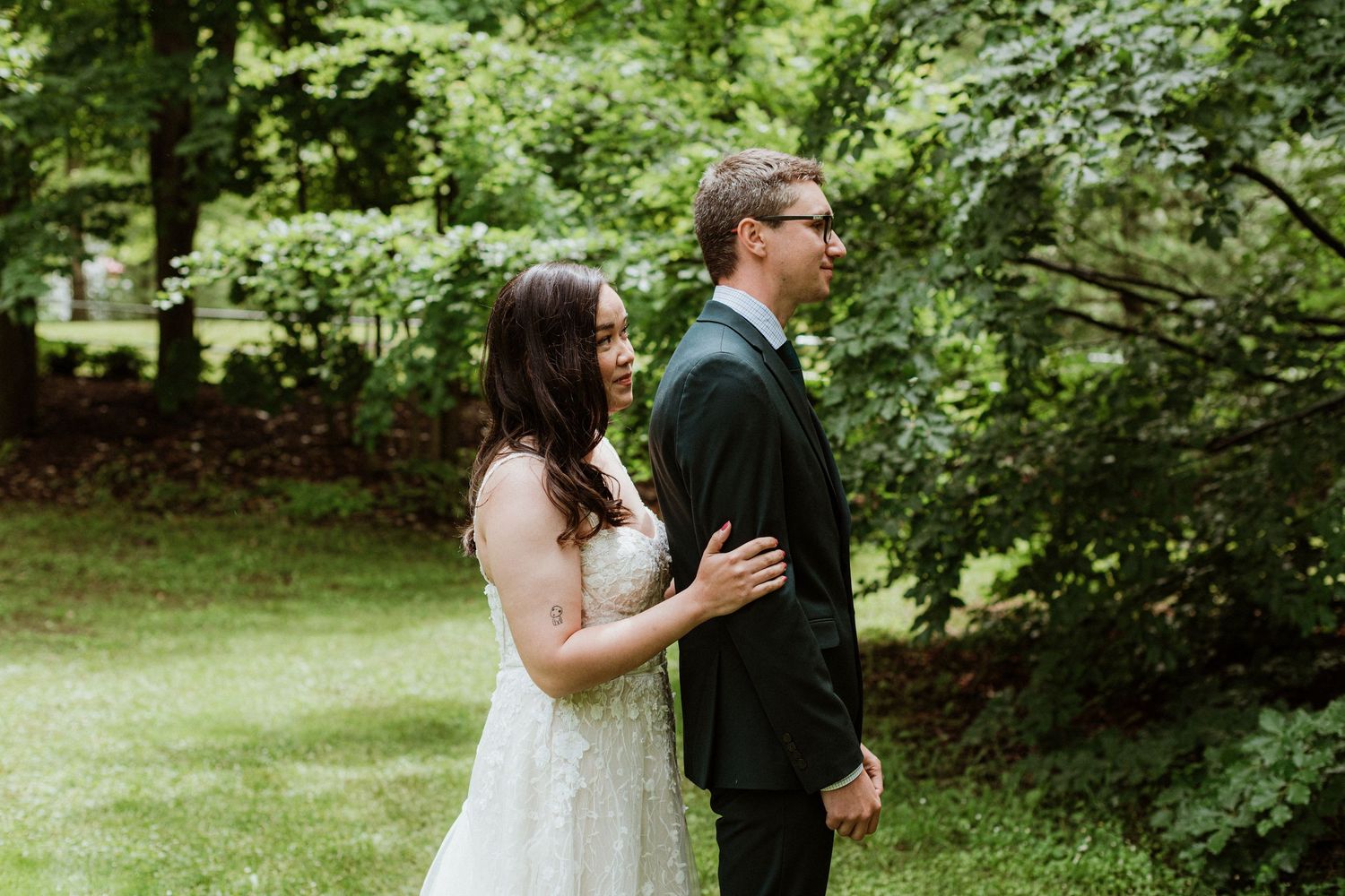 Bride in lace gown approaching groom from behind for first look in lush garden.