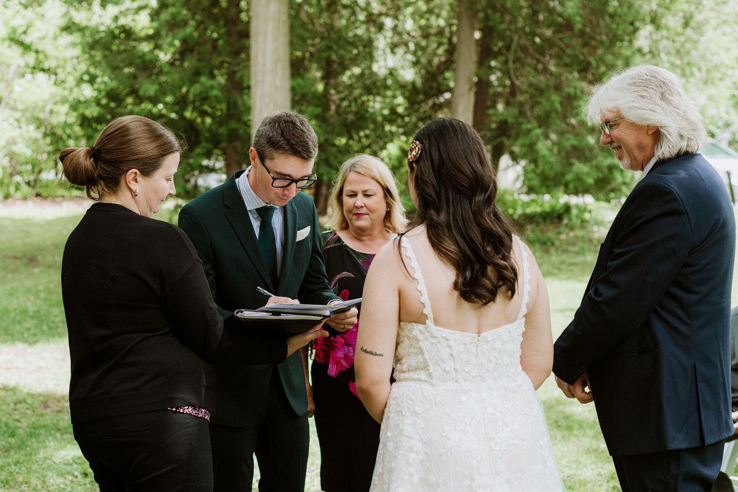 Wedding ceremony outdoors with officiant, bride, groom, and guests gathered in a lush green garden setting.