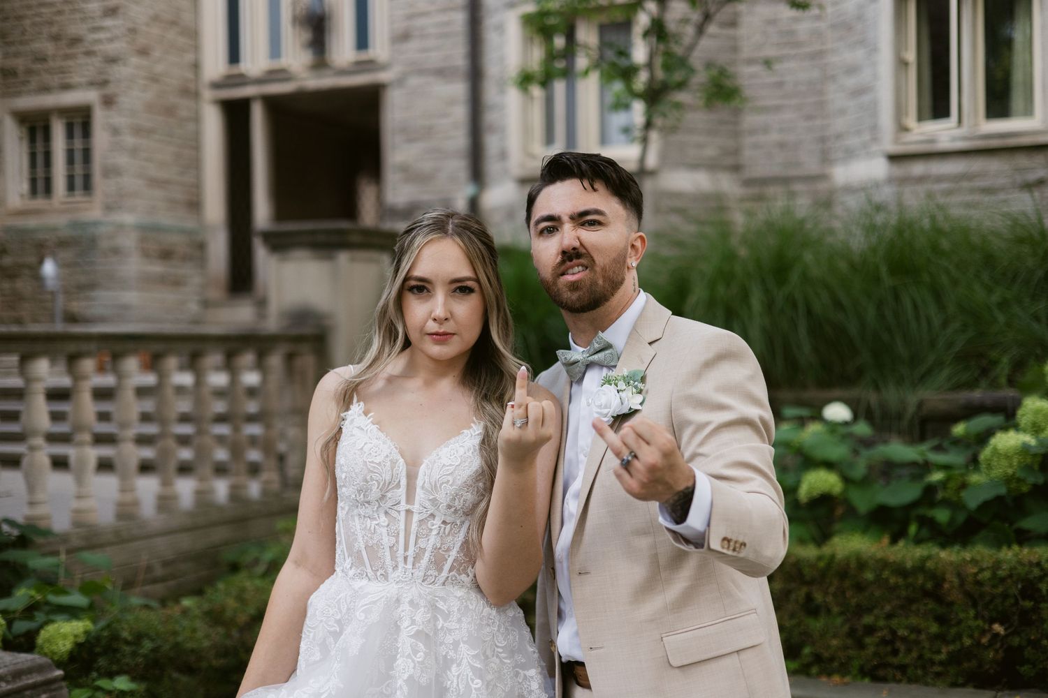 Bride in lace gown and groom in beige suit make playful faces outside stone venue.