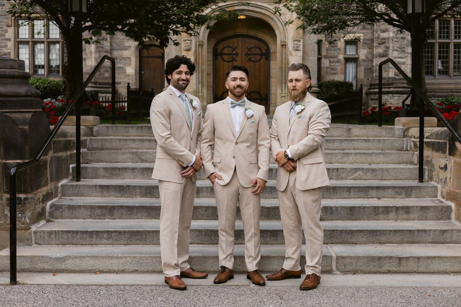 Groom and two groomsmen smile in matching tan suits on stone steps outside historic stone building.
