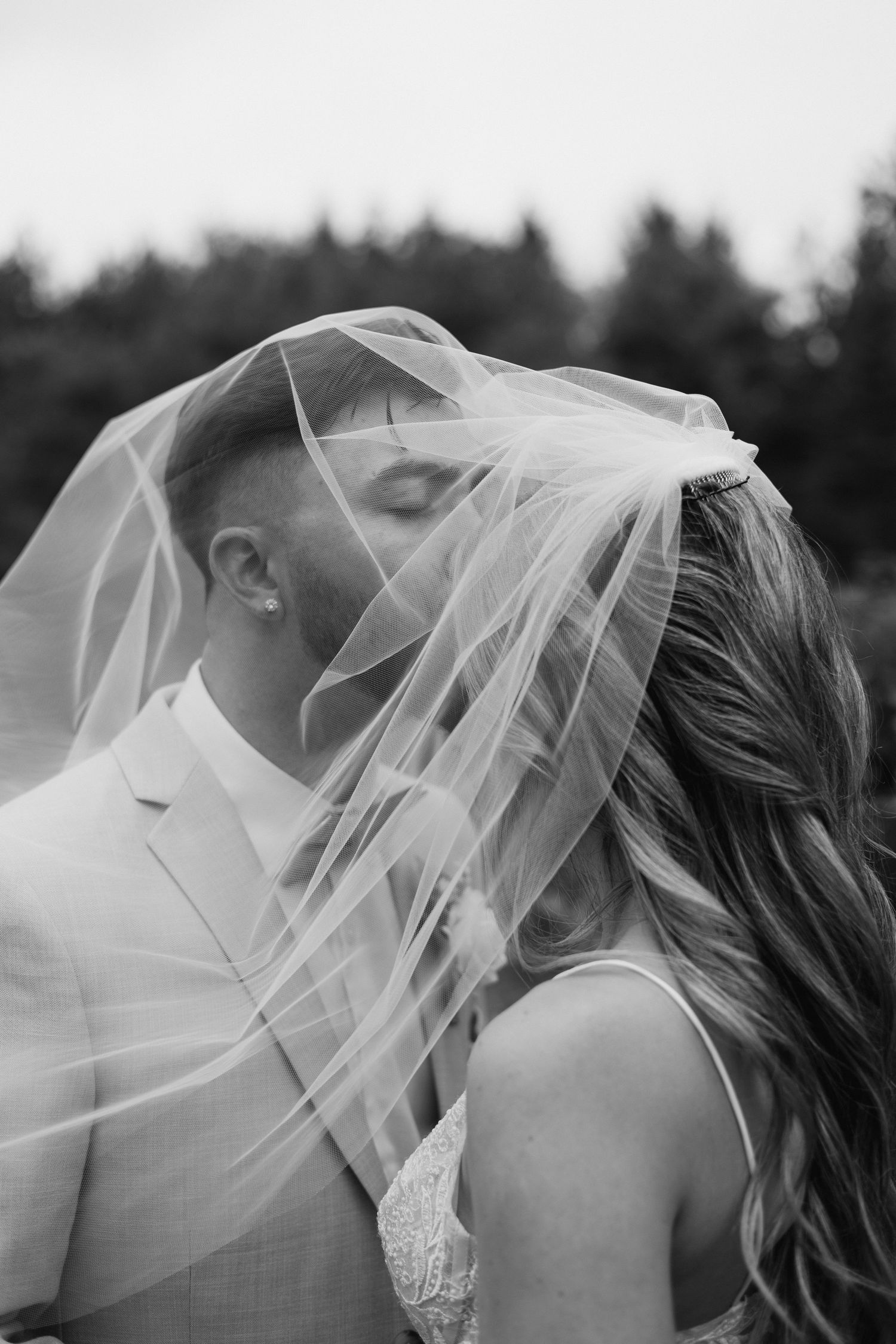 Black and white photo of bride and groom kissing under billowing veil with forest backdrop.