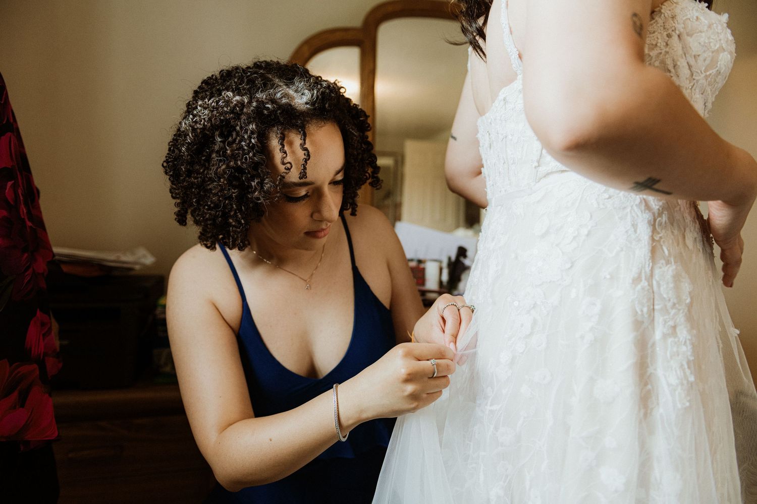 Bridesmaid in navy dress carefully buttoning the lace wedding gown of the bride before ceremony.