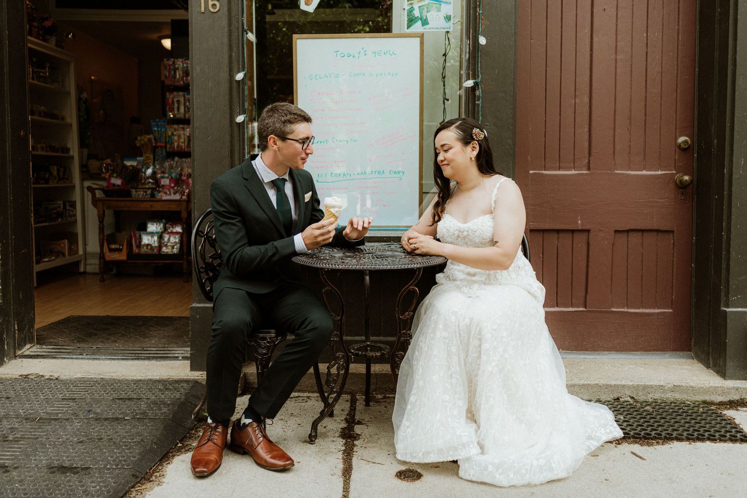 Bride and groom sitting at an outdoor cafe table sharing ice cream in wedding attire.