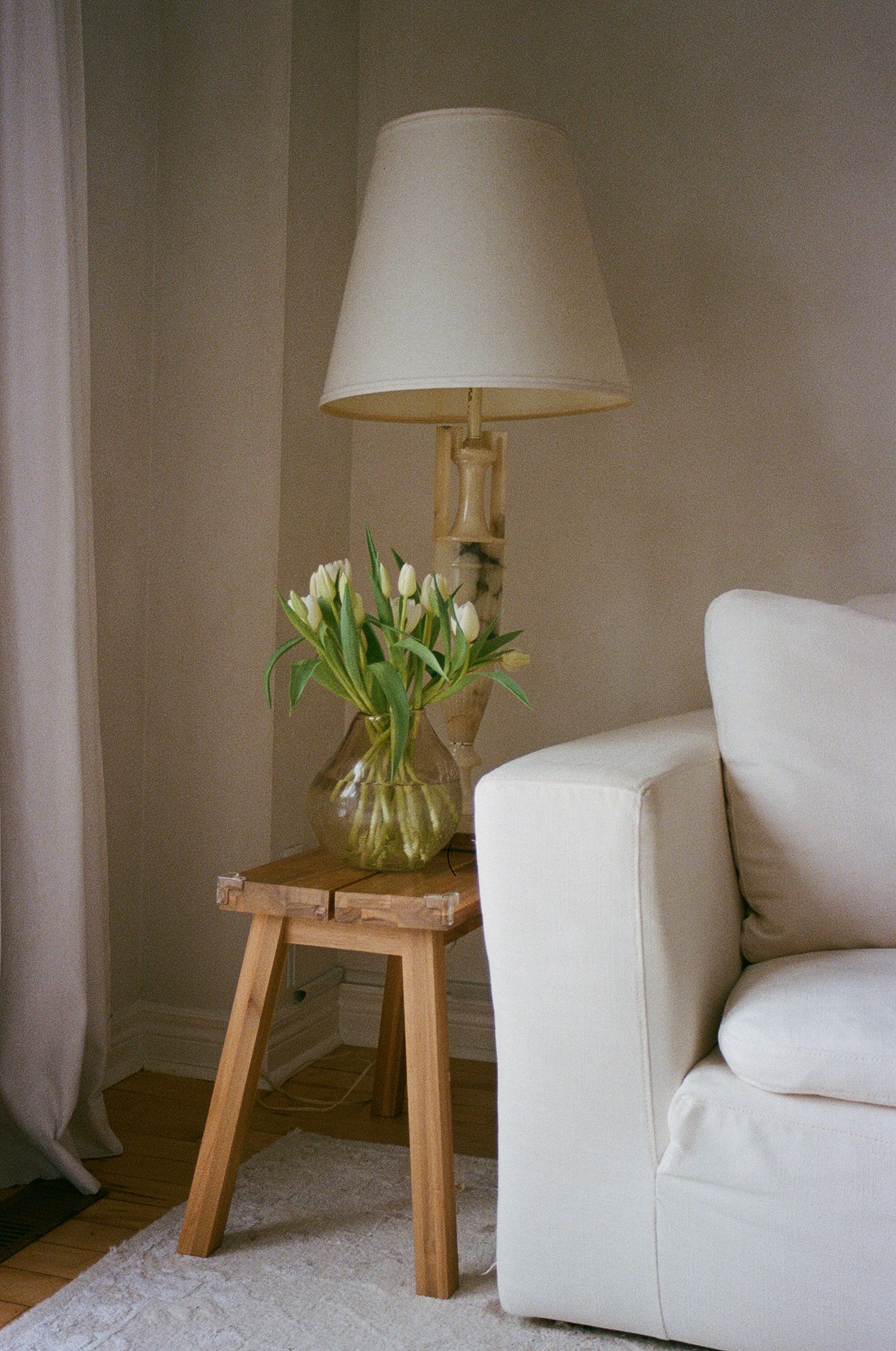 Minimalist wooden side table with white tulips in glass vase beside floor lamp and sofa.