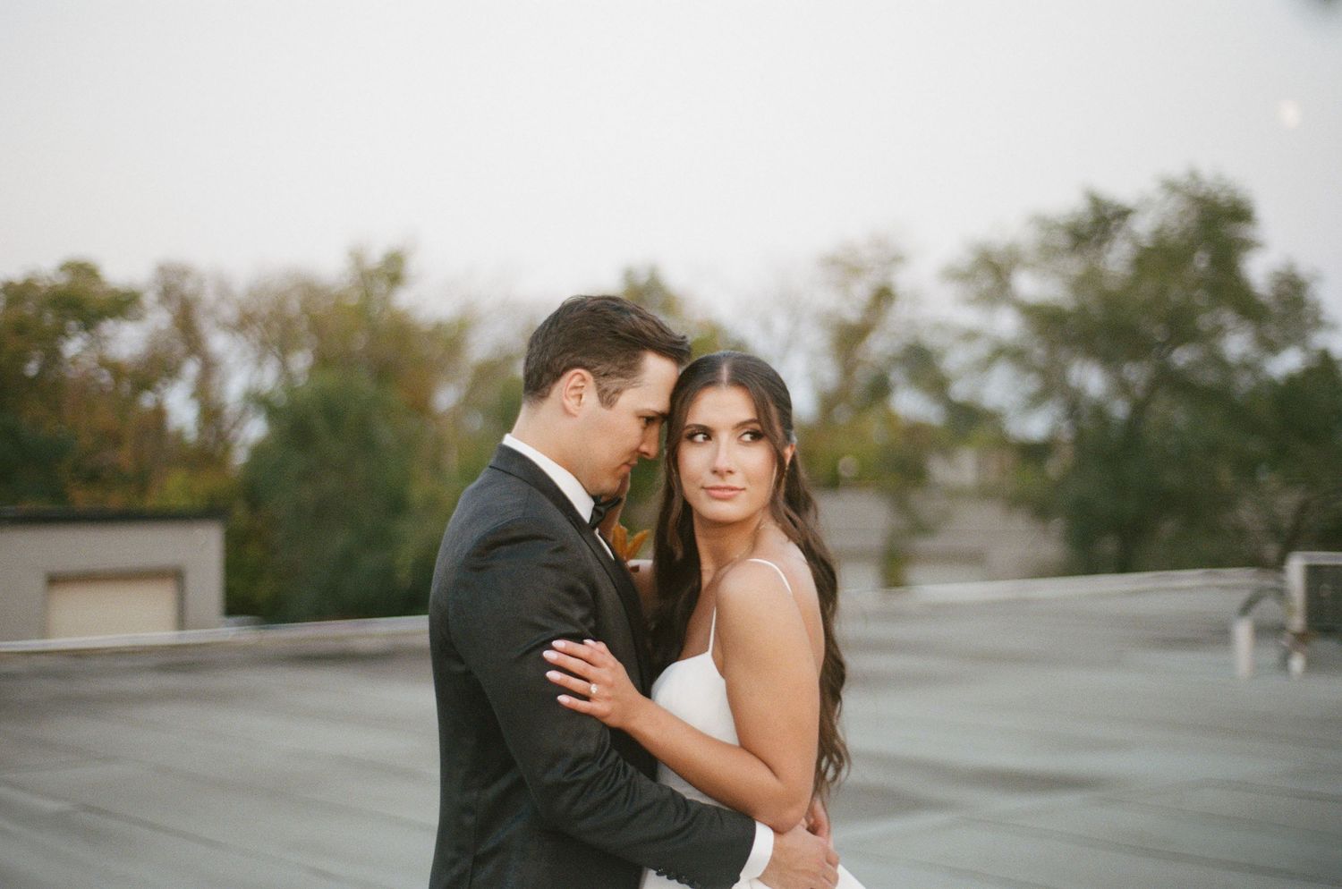 Bride looks directly at camera while groom nuzzles her in close romantic rooftop wedding portrait.