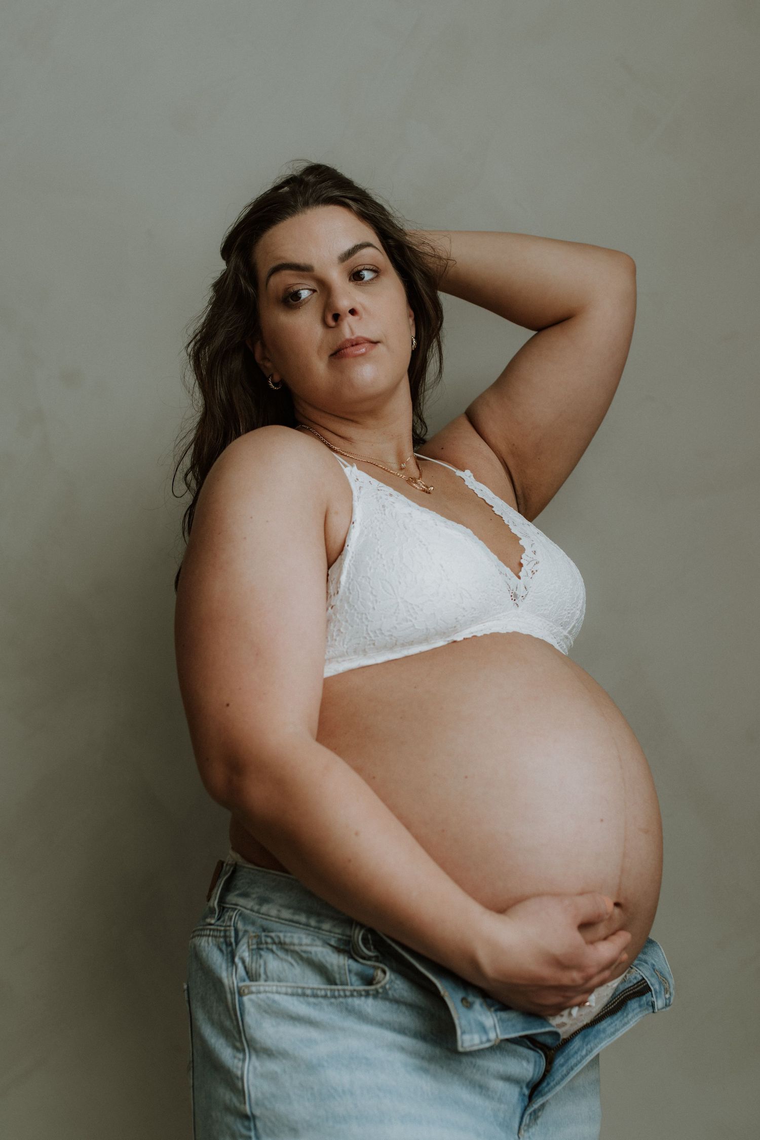 Pregnant woman in white lace bra and open jeans posing confidently, showcasing her baby bump.