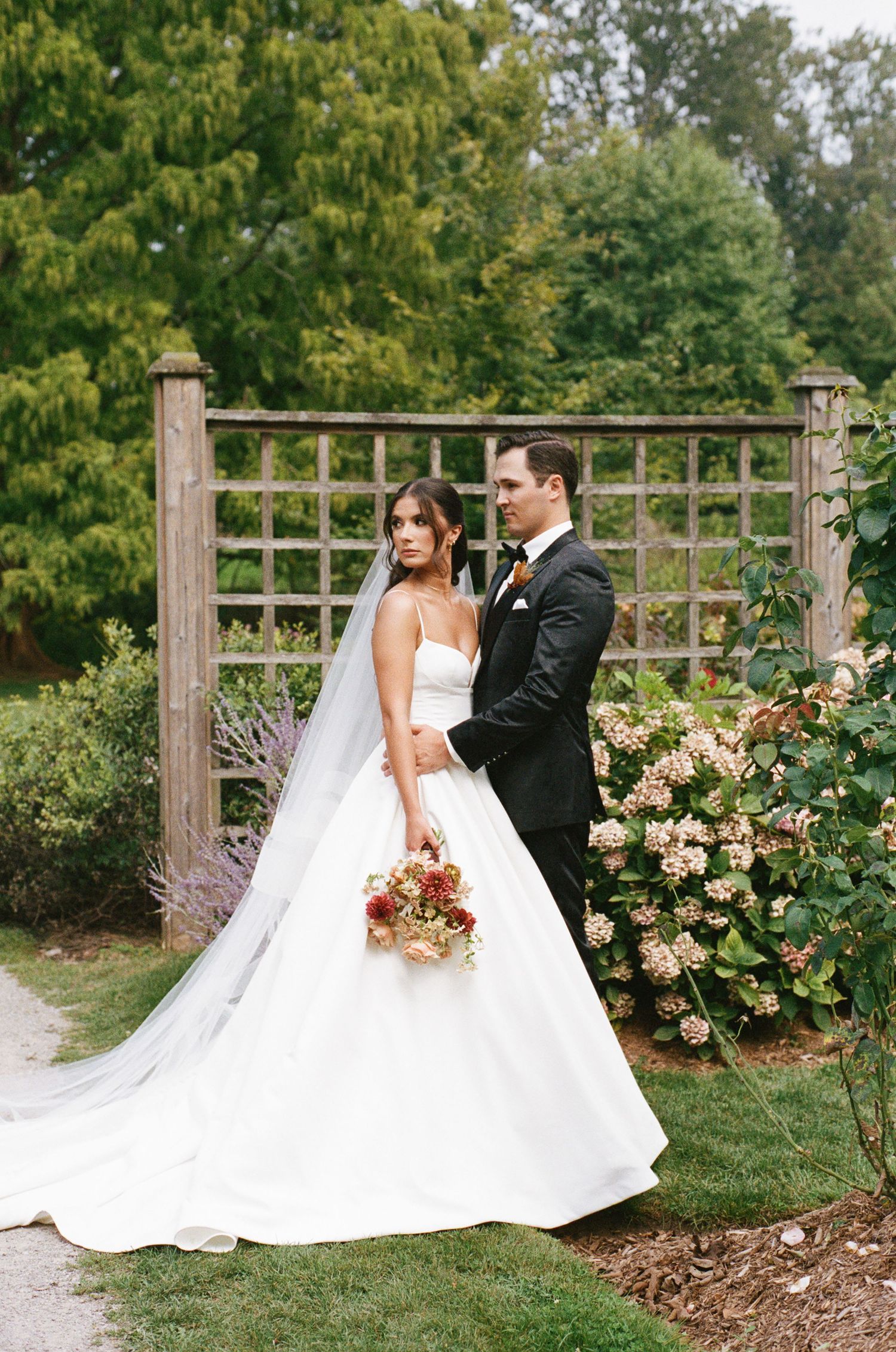 A bride in a strapless ballgown with a long veil holds a bouquet beside her tuxedo-clad groom in a garden.