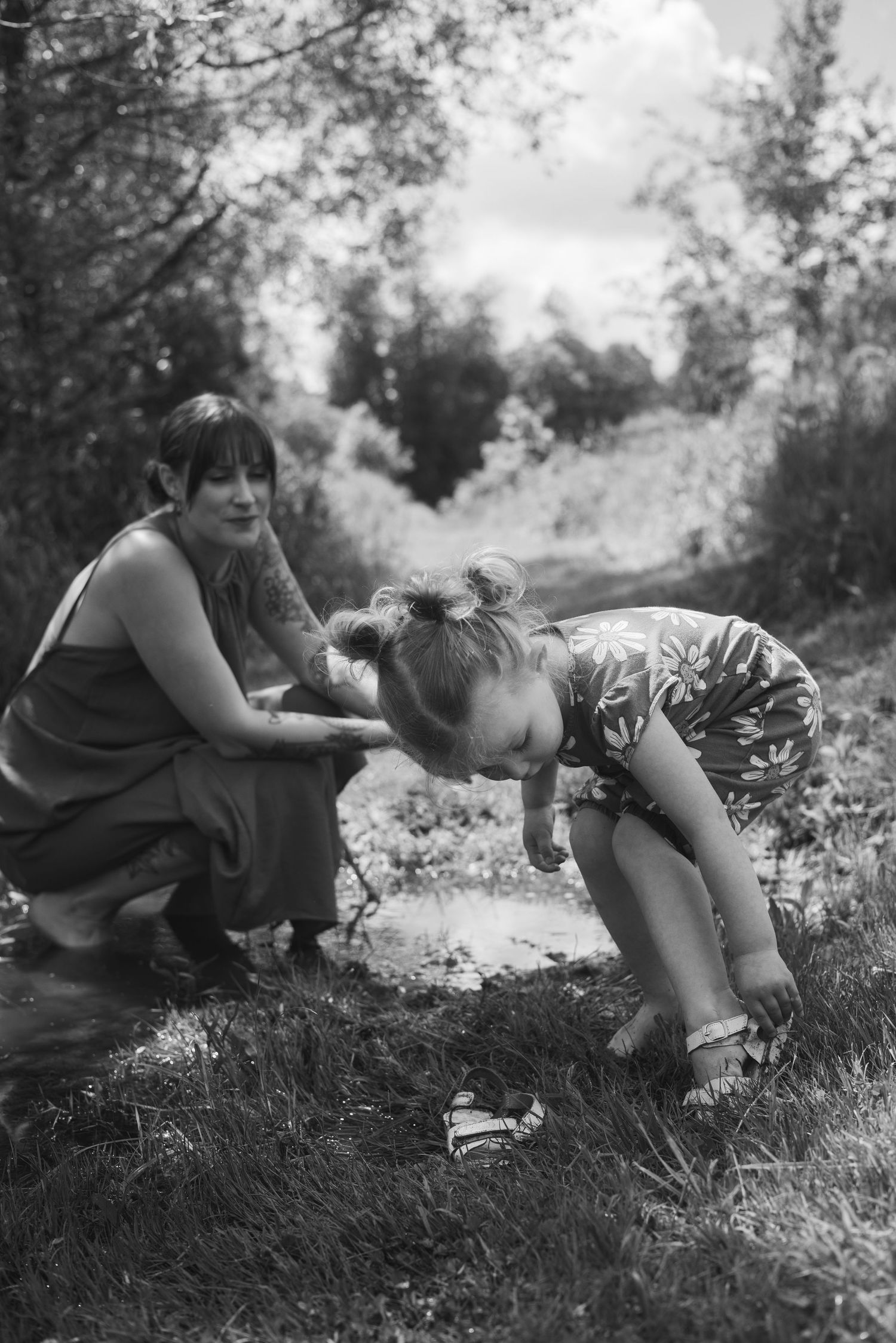 Black and white photo of a tattooed mother watching her young child bend down to explore a small stream outside.