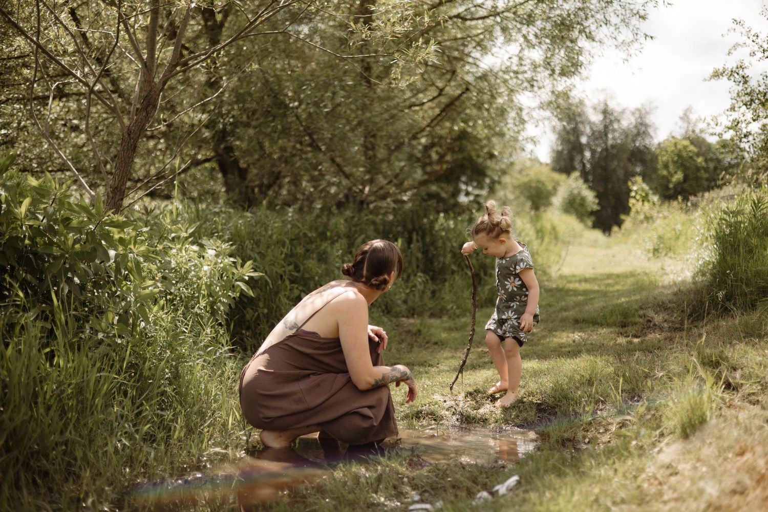 Mother crouches by a stream as young child plays with a stick in a lush green outdoor setting.