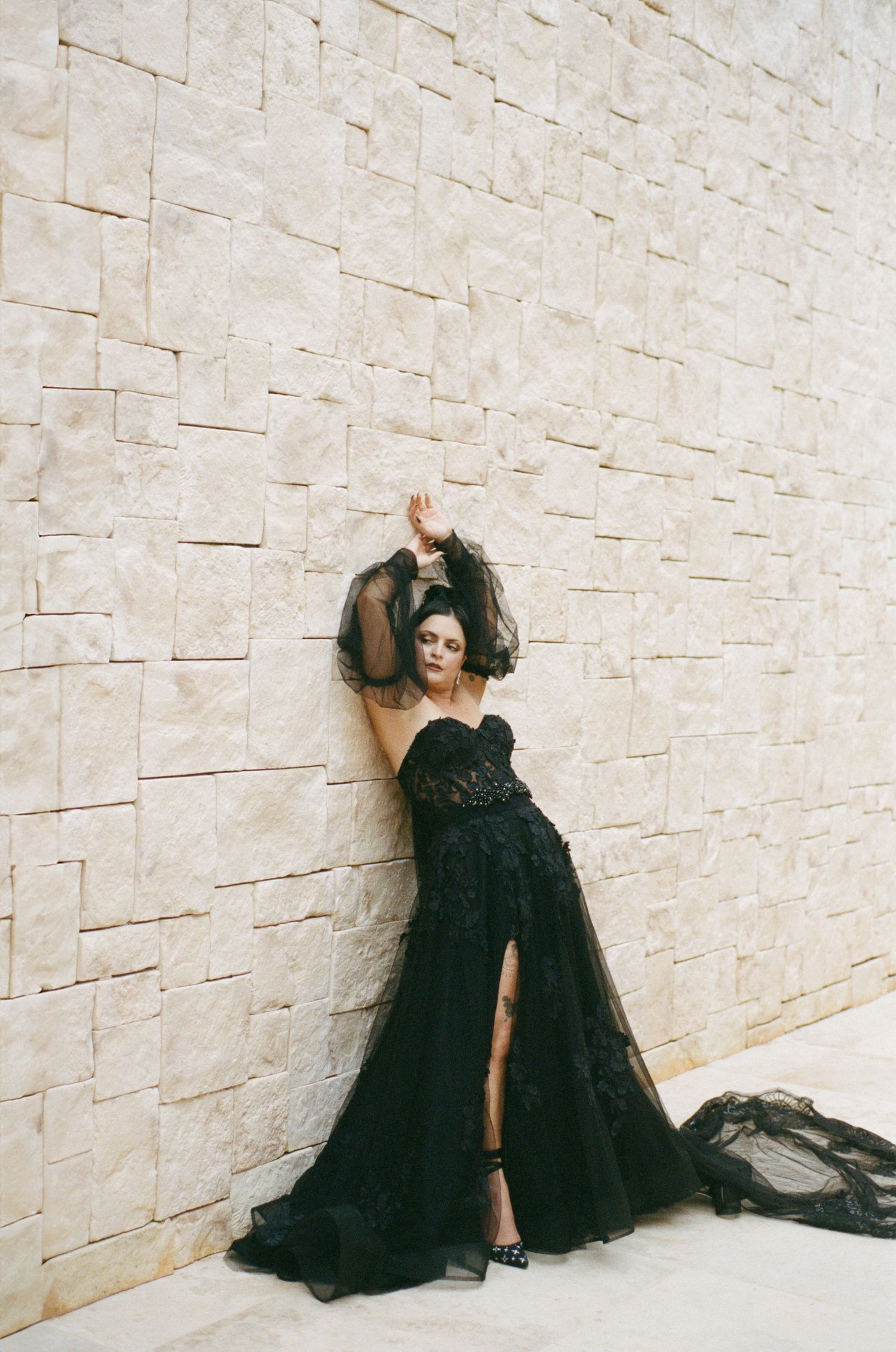 Bride in black strapless lace gown with tulle skirt posing dramatically against stone wall.