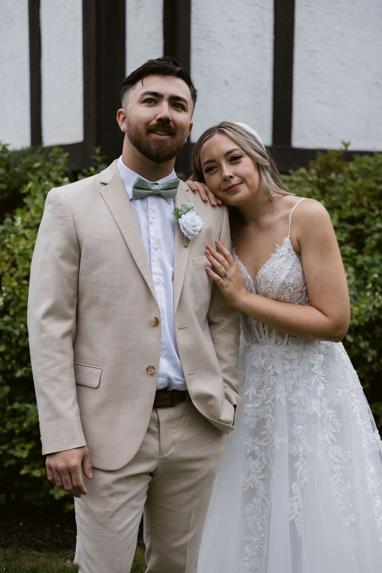 Bride and groom posing together outdoors, groom in beige suit with bow tie, bride in lace wedding dress.