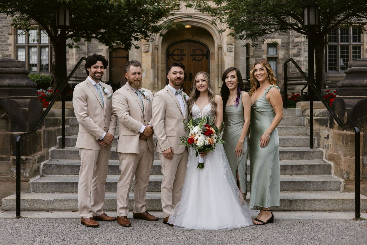 Wedding party poses on stone steps, groomsmen in tan suits and bridesmaids in sage green gowns with bride.
