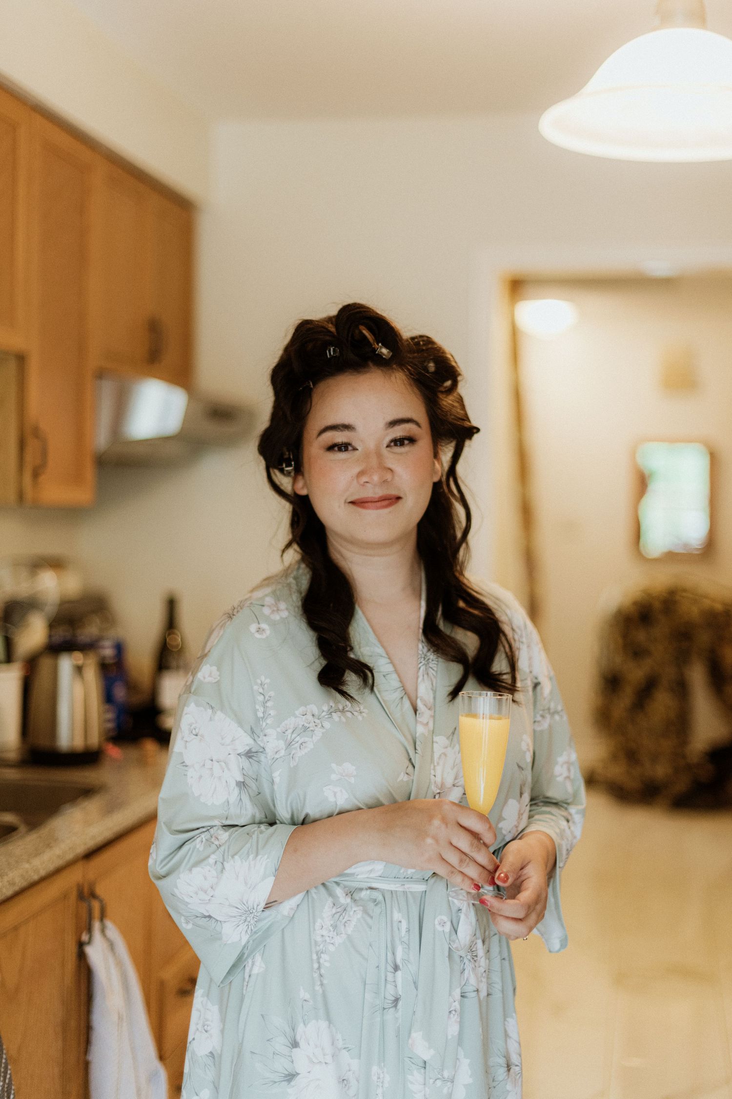 Bride in a floral robe with hair in rollers holds a champagne flute while smiling in a warm kitchen.