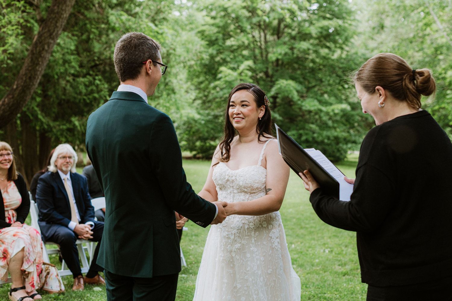 Couple holds hands and smiles during outdoor wedding ceremony while officiant reads from black book.