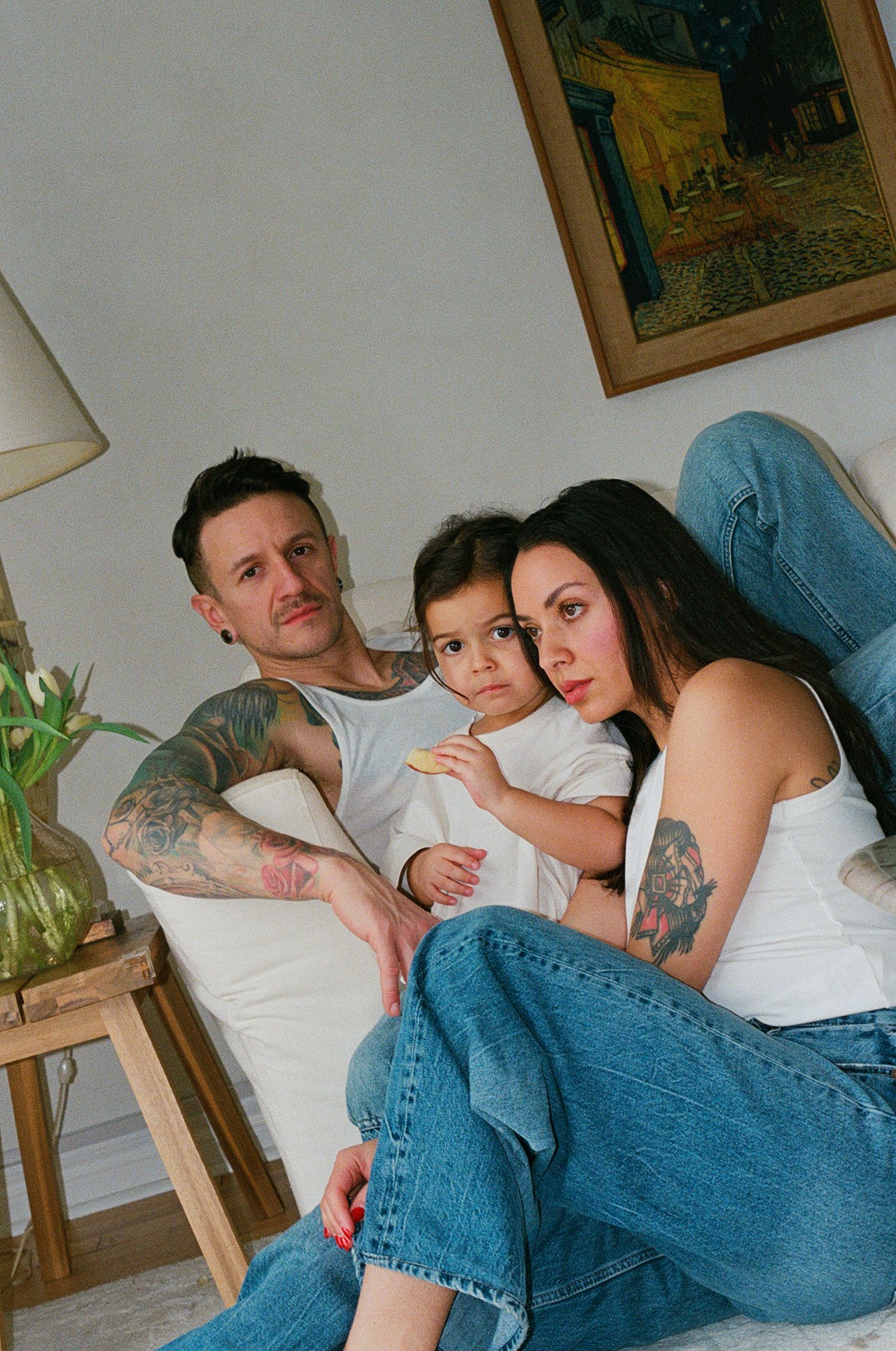 Tattooed couple with young child relaxing on white sofa, posing for family portrait at home.