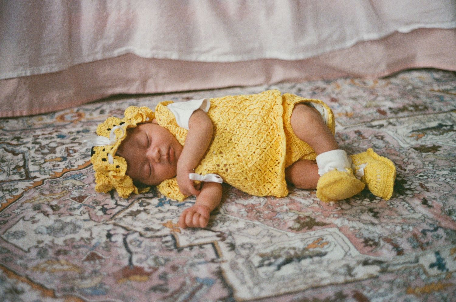 Sleeping newborn baby dressed in a yellow crocheted outfit lying on a floral rug.