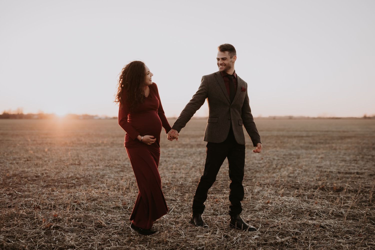 Expecting couple holds hands walking through golden sunlit open field at sunset.