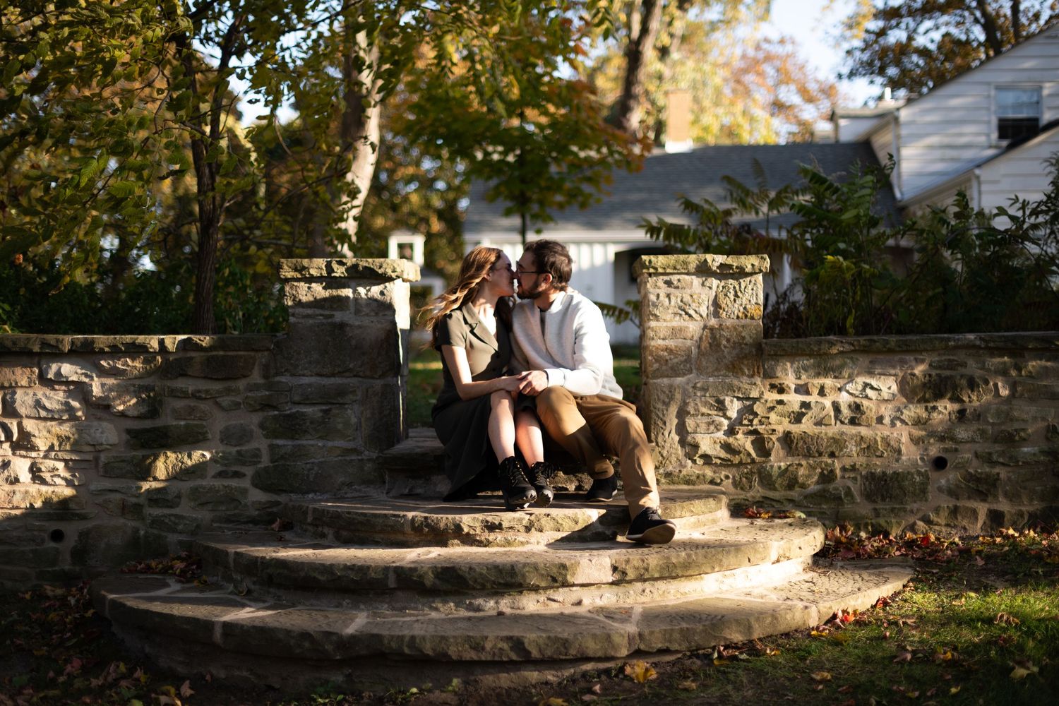 Couple sharing a kiss while sitting on rustic stone steps surrounded by autumn foliage.