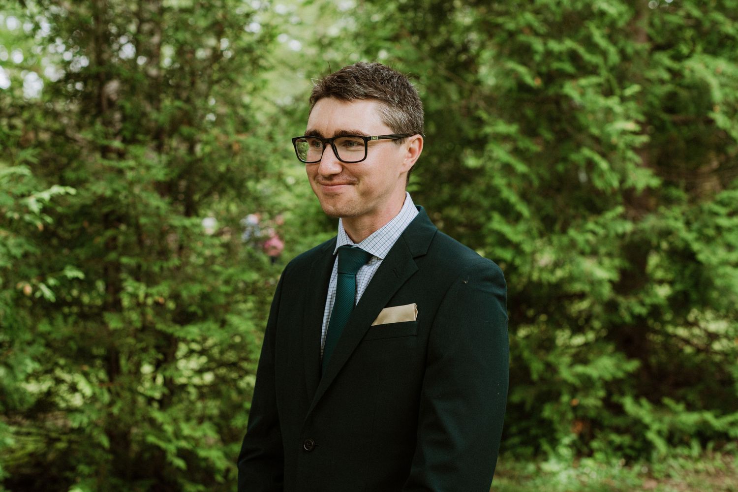 Dapper groom in dark suit and glasses smiles confidently outdoors surrounded by green trees.