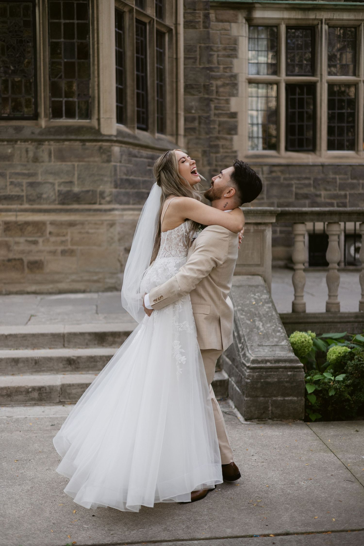 Groom lifts laughing bride in white tulle wedding gown outside a Gothic stone building.