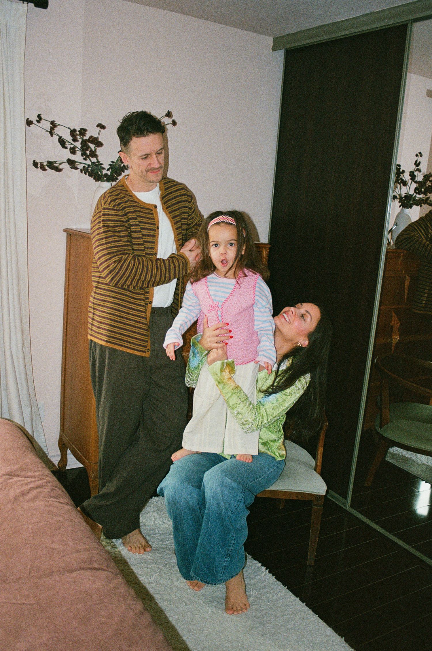 Family playing together in bedroom with father lifting young daughter while mother sits on chair.