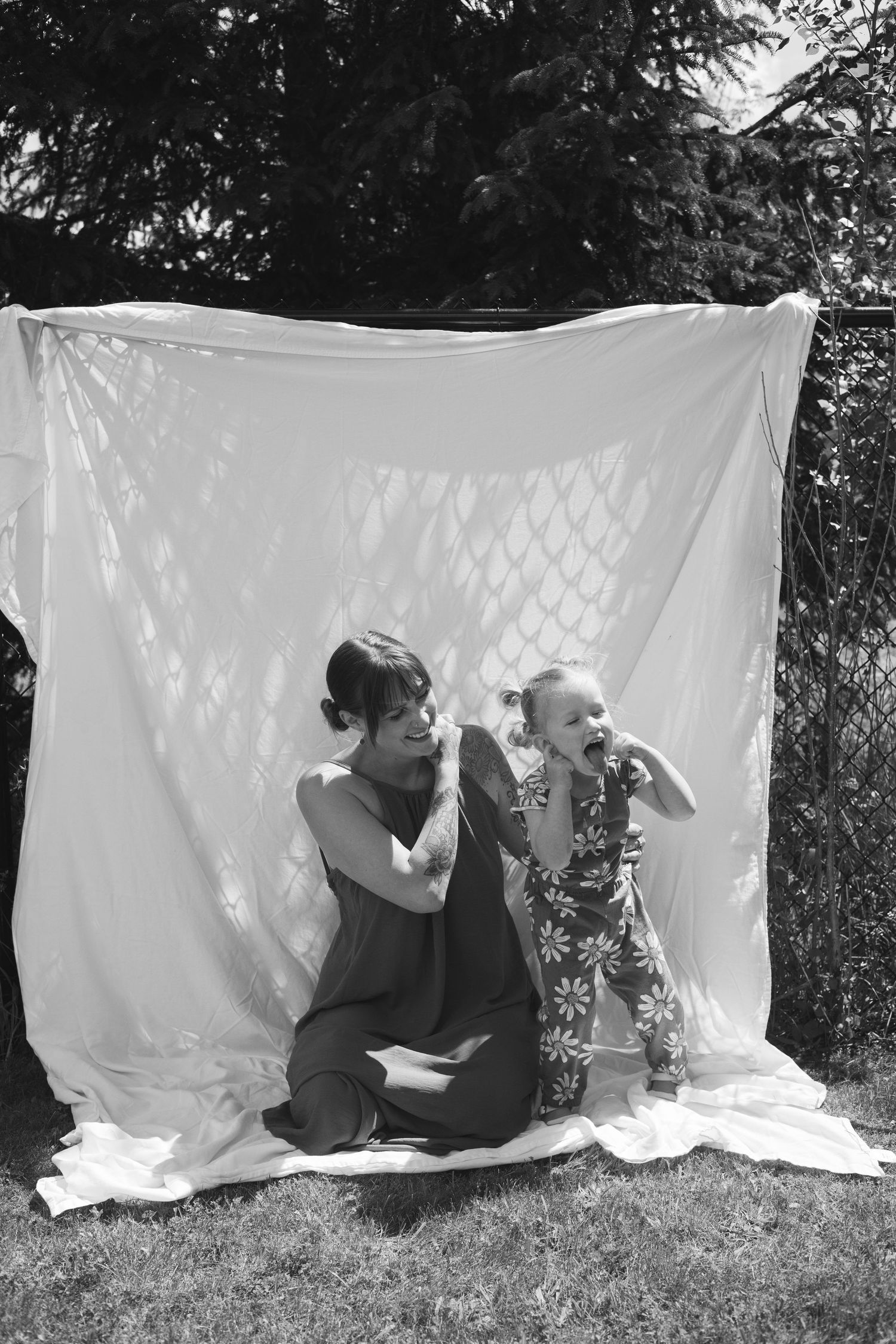 Black and white photo of mother and toddler daughter sitting together against a white sheet backdrop in backyard.