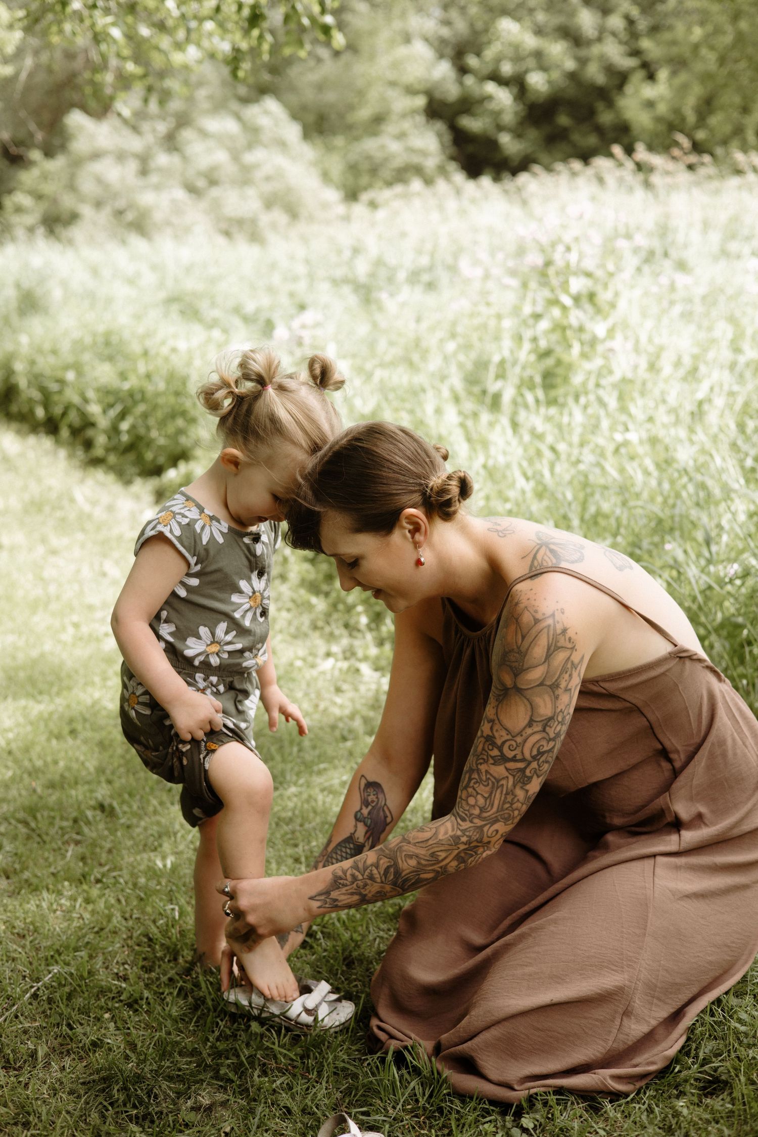 A tattooed mother kneels on grass to help her young daughter put on sandals in a lush green outdoor setting.