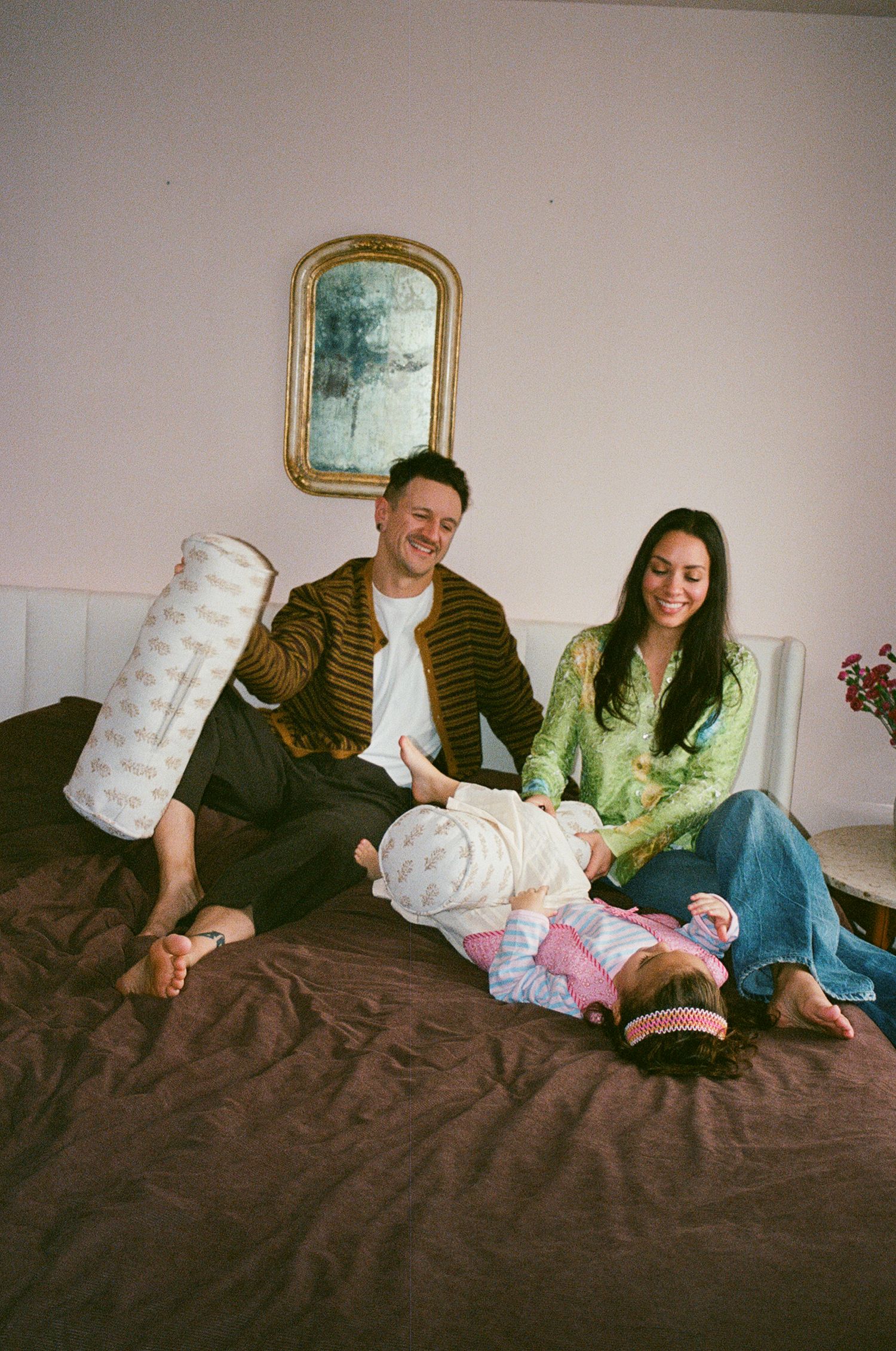 Happy couple sits on bed with baby lying between them, man holding decorative pillow, gold mirror behind.