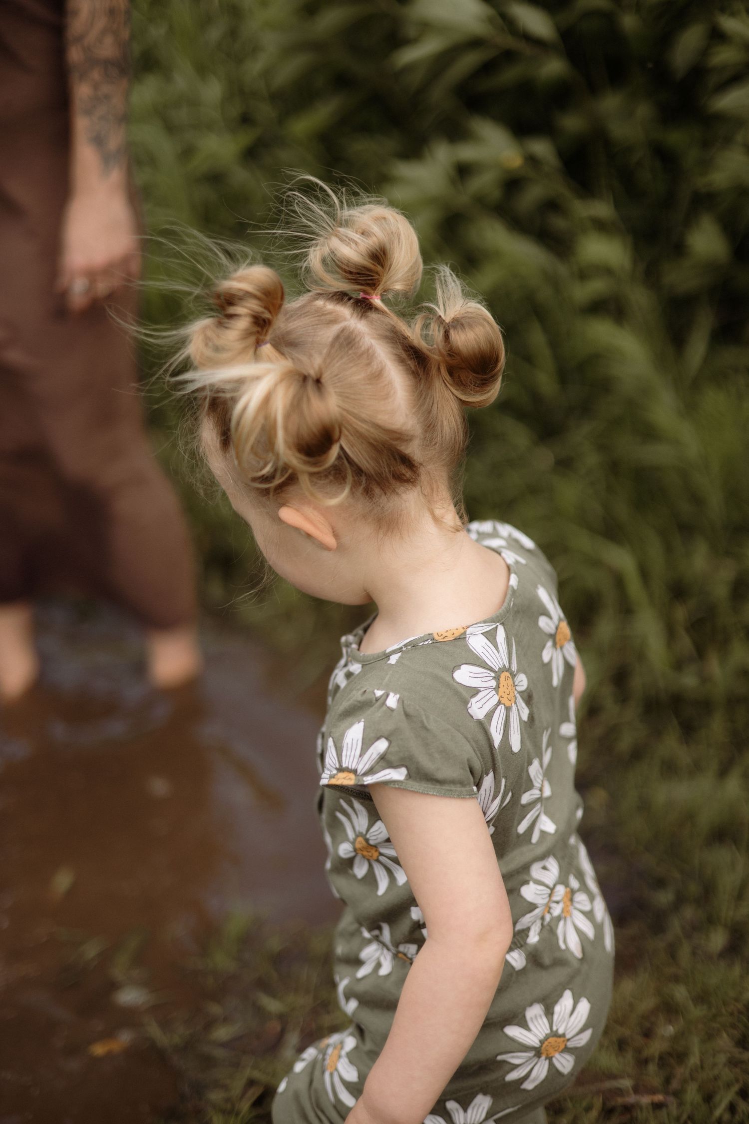 Close-up of toddler girl with double bun hairstyle wearing green daisy print romper outdoors.