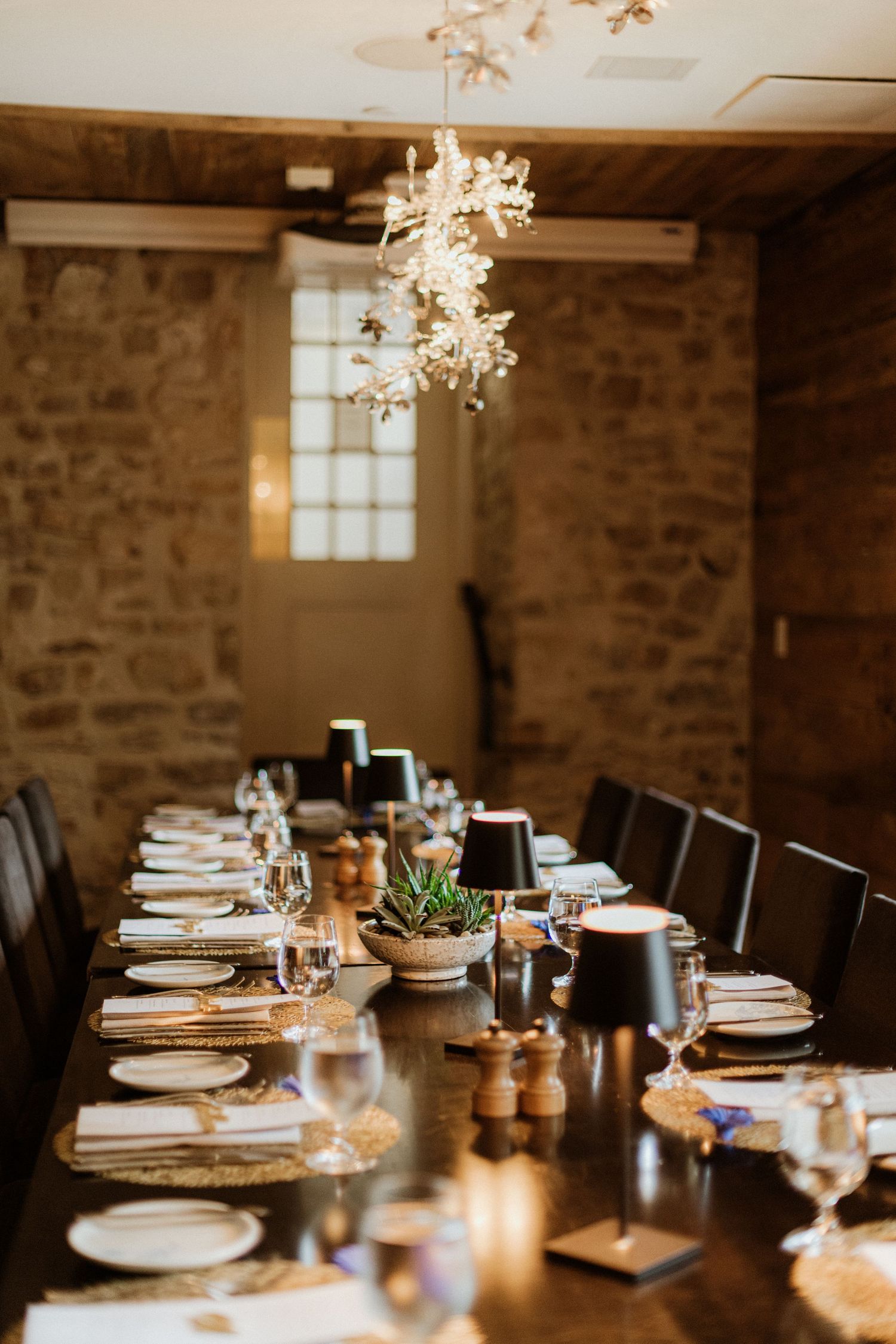 Elegant long dining table set for wedding reception in rustic stone room with chandelier.