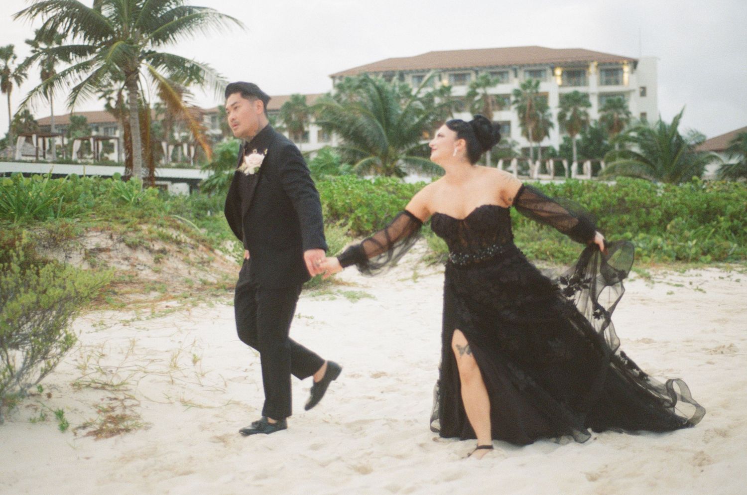 Couple in matching black wedding attire hold hands walking on sandy beach.